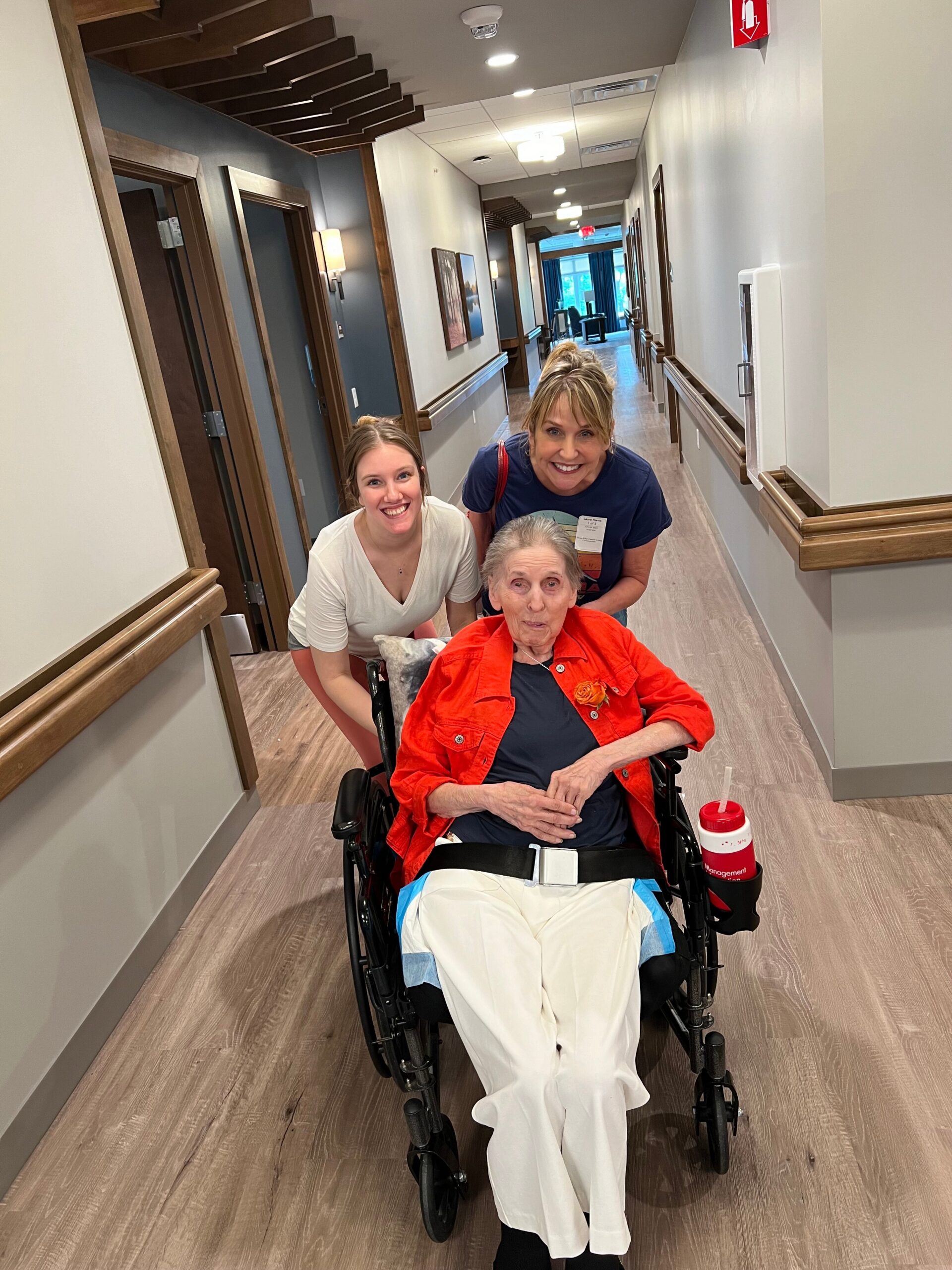 Elderly woman in a wheelchair flanked by two smiling caregivers in a hospital hallway.