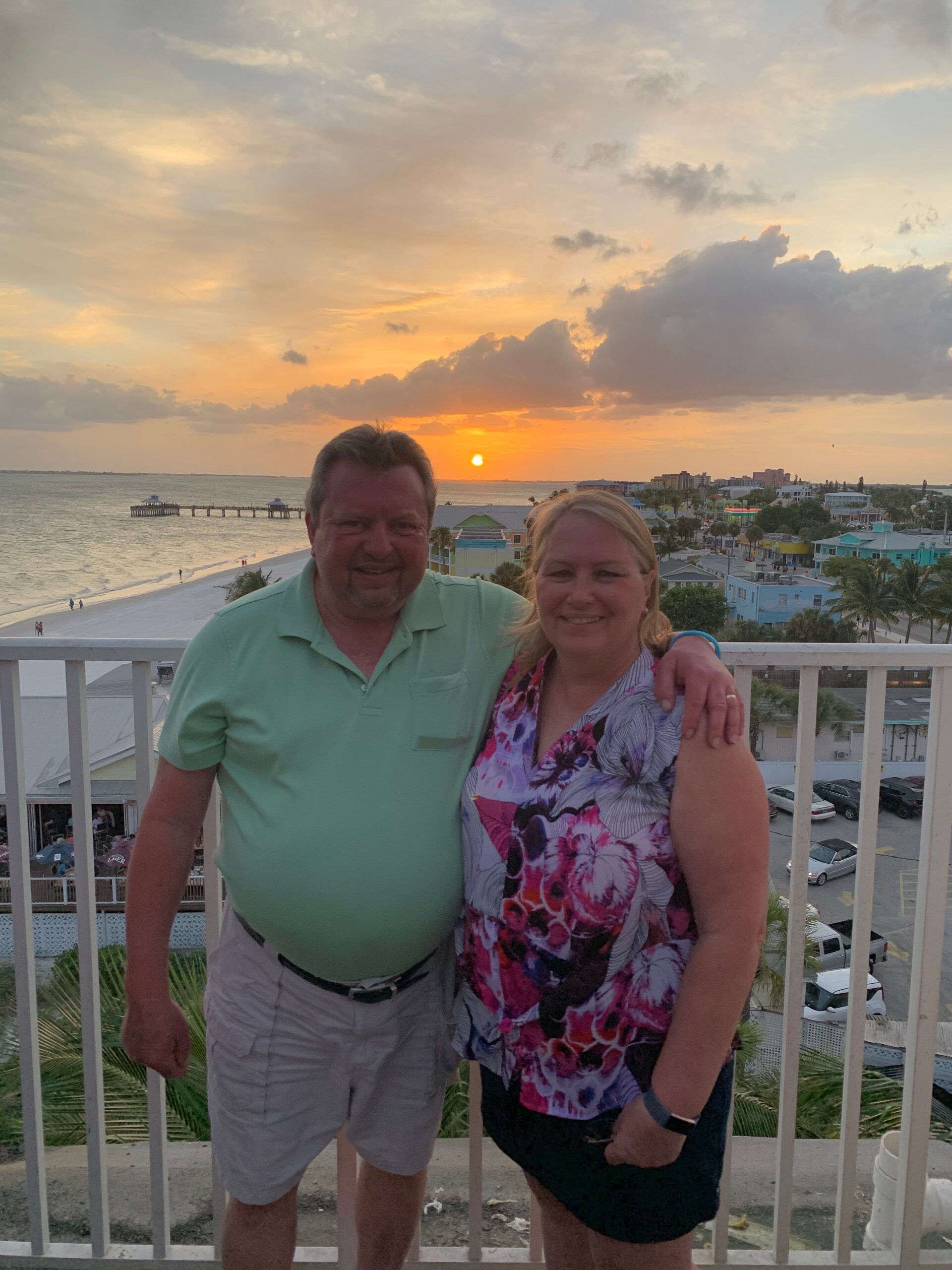 Smiling couple on a balcony at sunset with the beach and pier in view behind them.