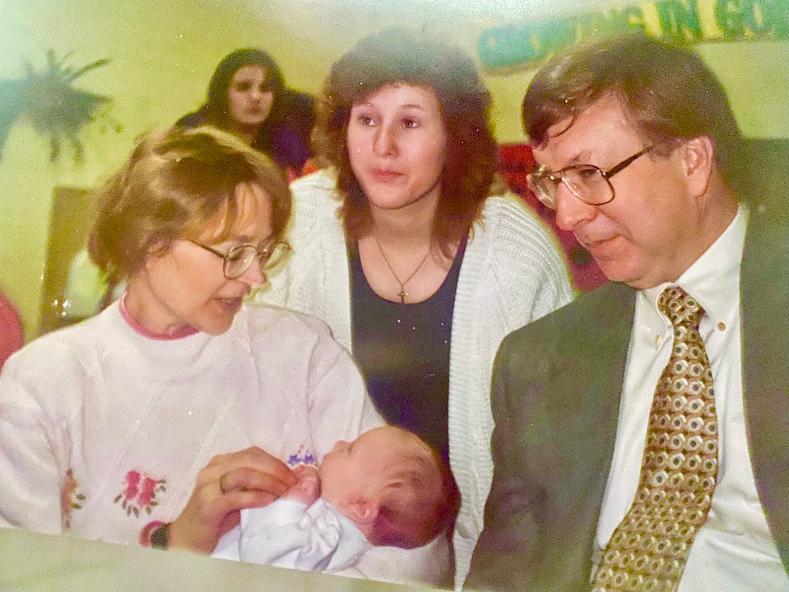 Newborn infant held by a woman in a light pink sweater, surrounded by family members in a warm gathering.