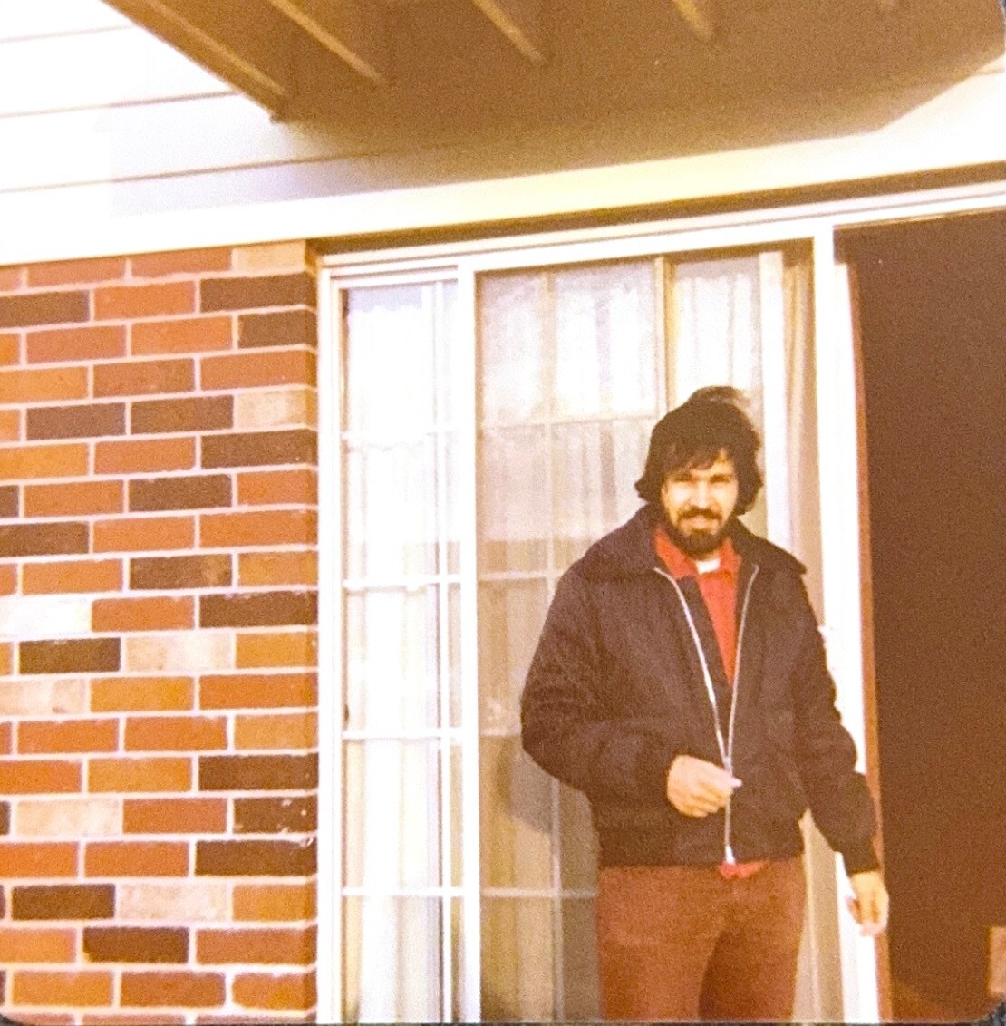 Man with dark hair and beard stands on a brick porch next to a glass door, wearing a dark jacket and red shirt.