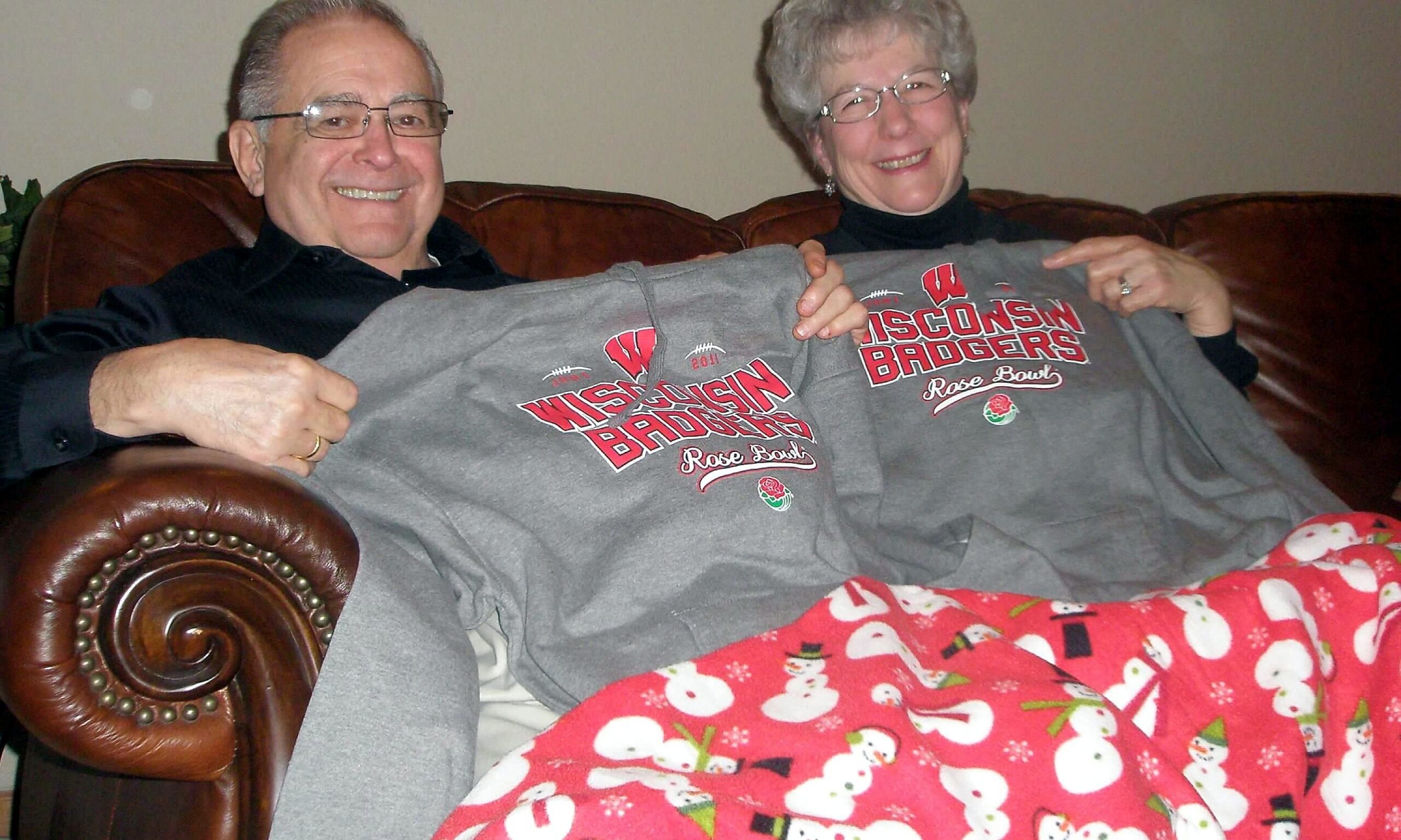 Smiling older couple sitting on a brown leather couch, proudly displaying matching gray Wisconsin Badgers Rose Bowl sweatshirts.