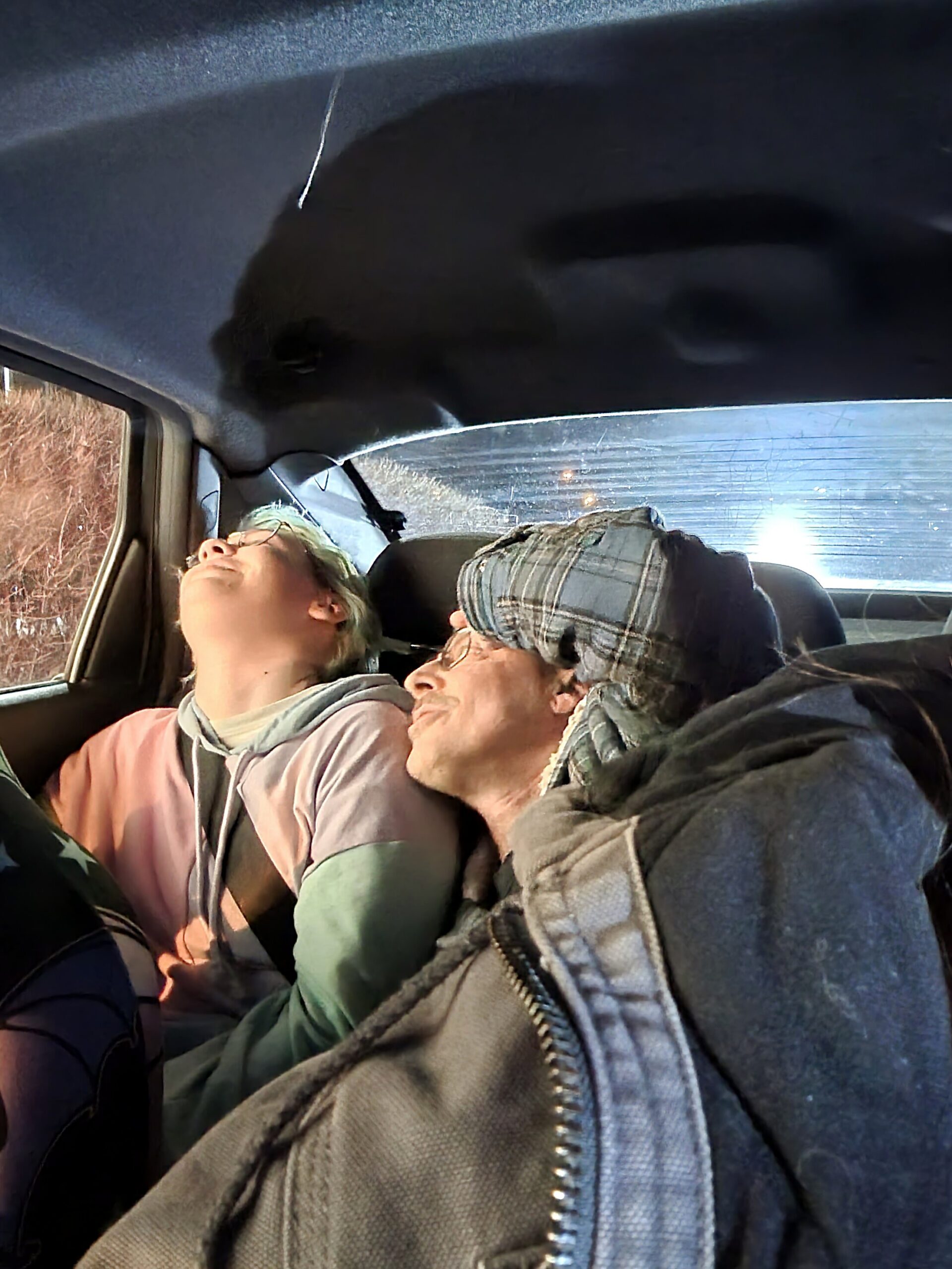 Two people riding in a car, leaning back and looking up at the ceiling; one person wears a pink-green hoodie while the other wears a plaid cap and scarf in the front seats.