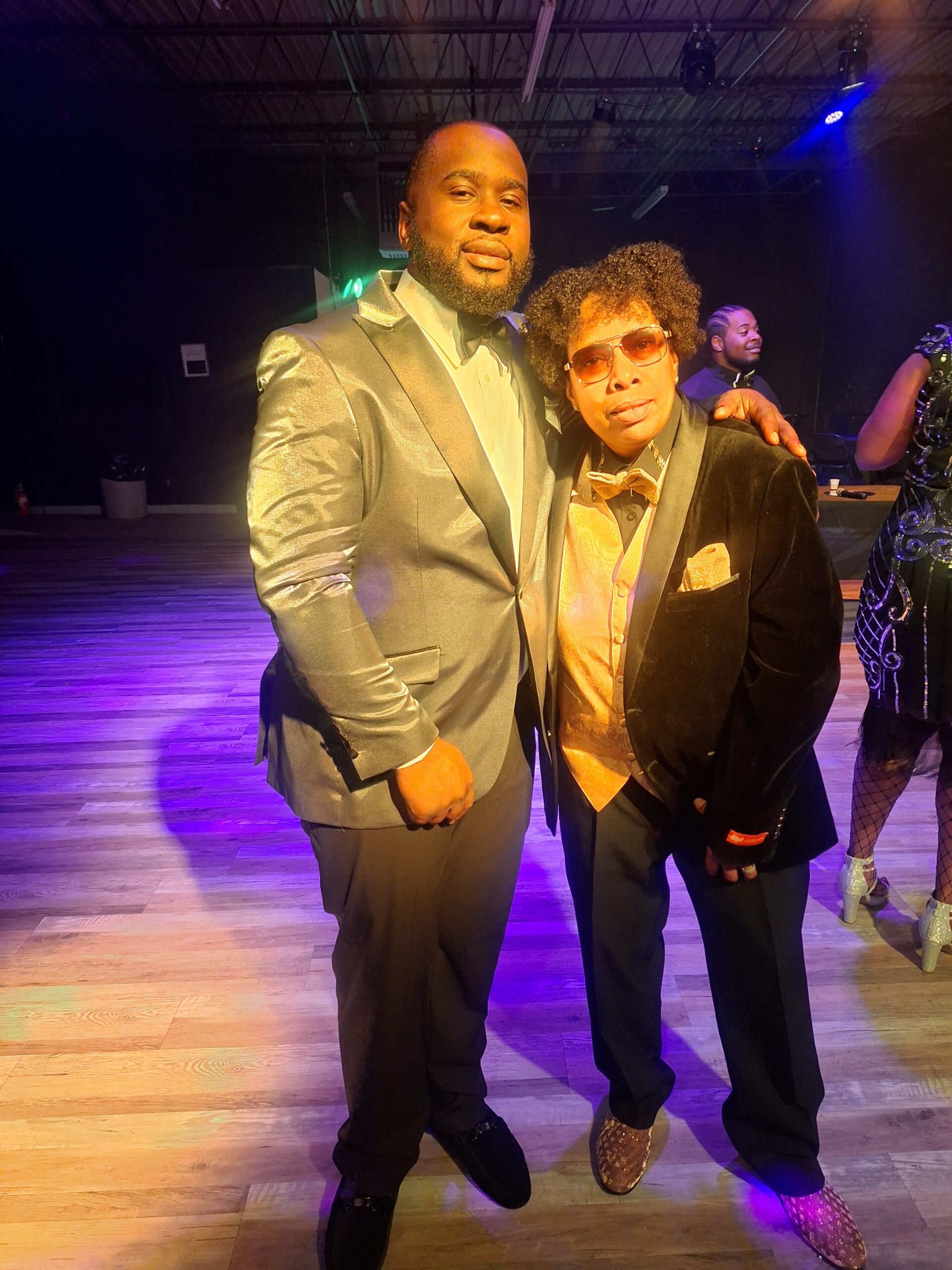 Two men in formal suits pose together on a dim-lit dance floor with colorful stage lighting behind them.