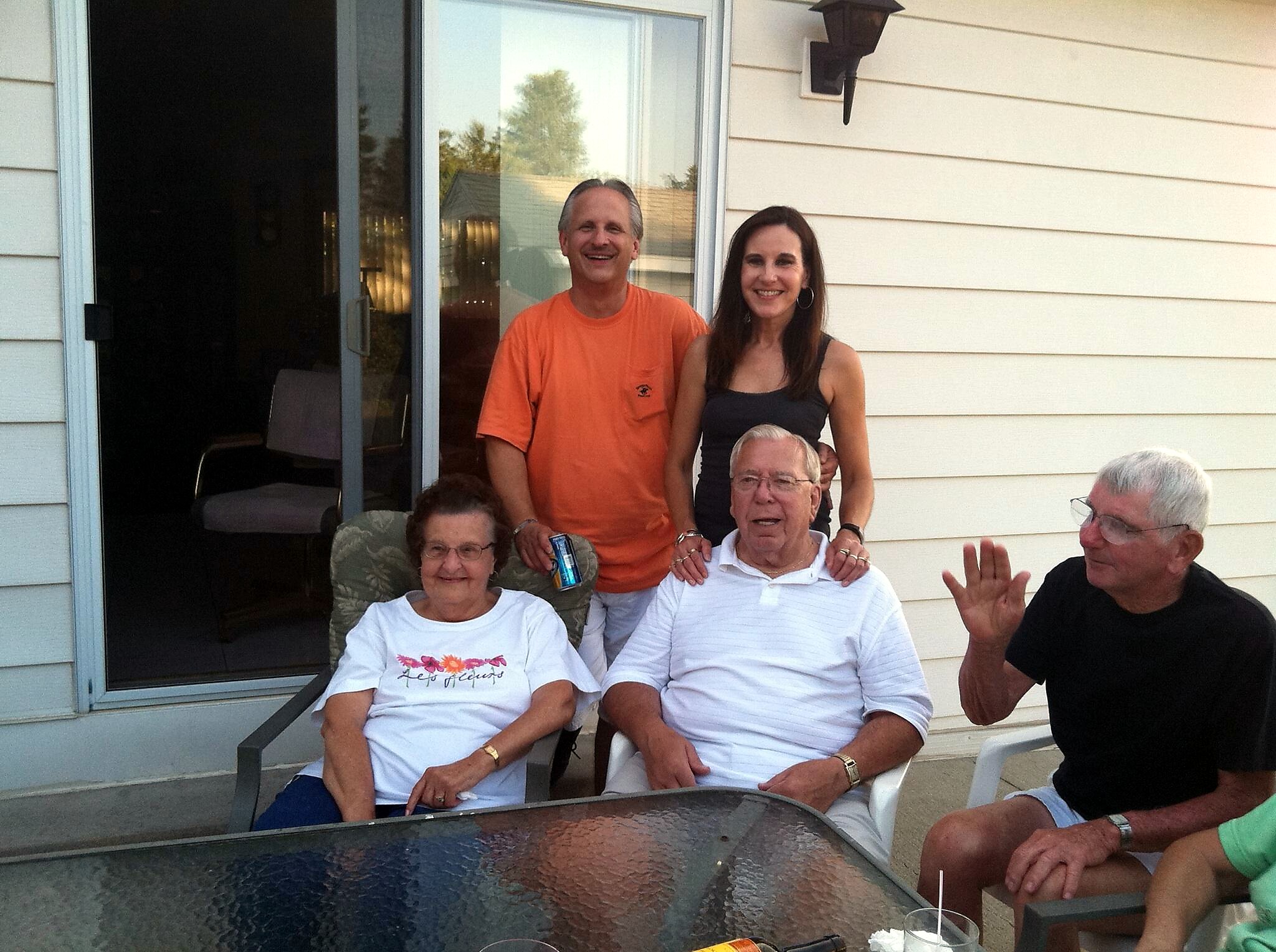A group of adults posing for a photo on a sunny patio outside a house, seated around a glass table with a sliding glass door in the background.