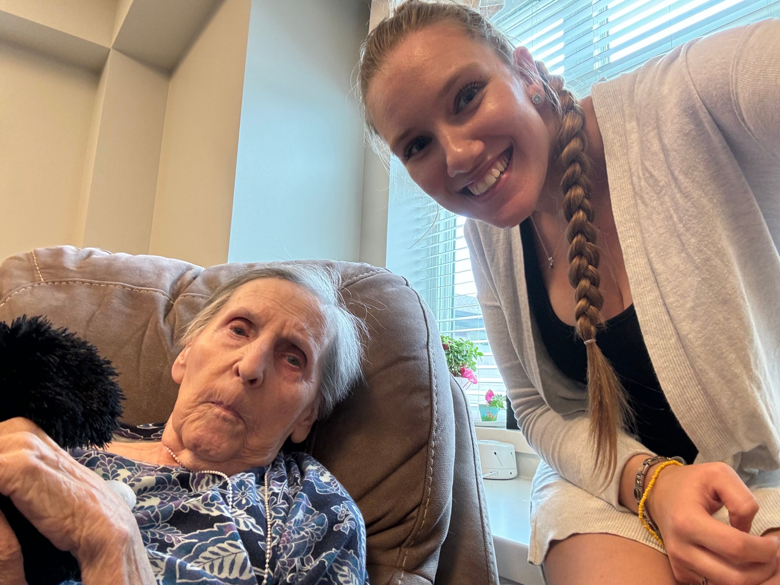 Two women taking a selfie indoors: an elderly woman seated on a beige couch wearing a blue floral top, and a smiling young woman with a long braid leaning in beside her near a window.