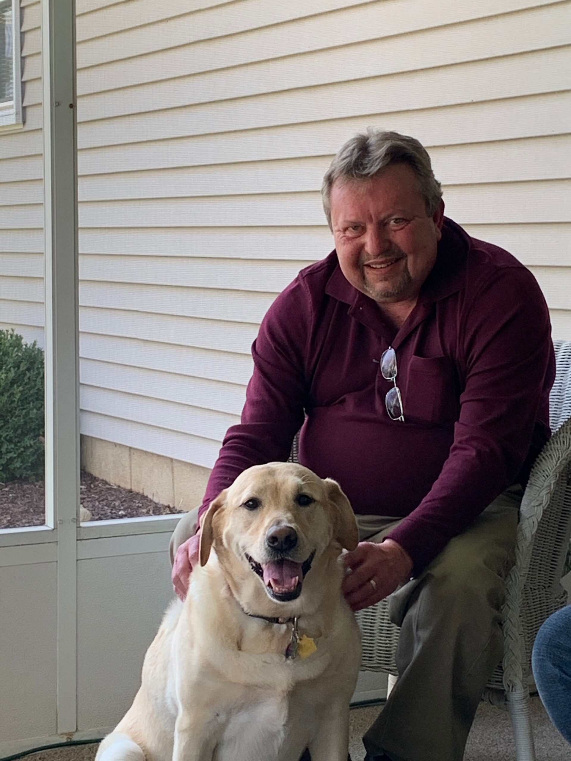 Smiling man in a maroon shirt sitting on a screened porch beside a large beige dog on a sunny day.