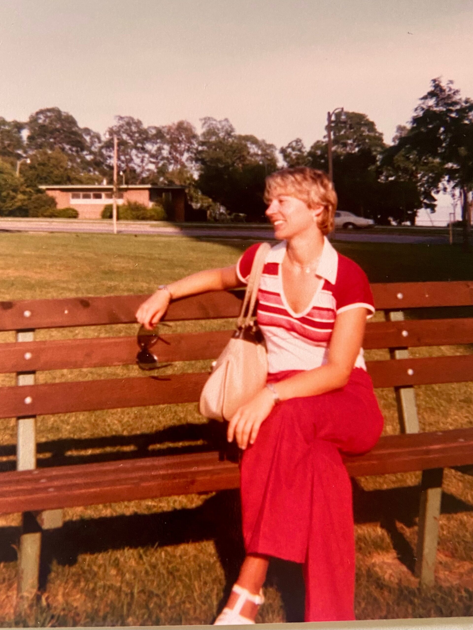 Woman in a red-and-white striped top and red skirt sits on a wooden park bench, smiling, with a beige bag over her shoulder on a sunny day in a park.