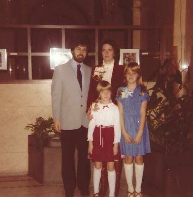 Family of four posing for a formal event: man in a light gray suit, woman in dark blazer with a corsage, and two young girls in dresses (one white top with red skirt, the other blue dress) standing indoors by a window with photos on the wall.