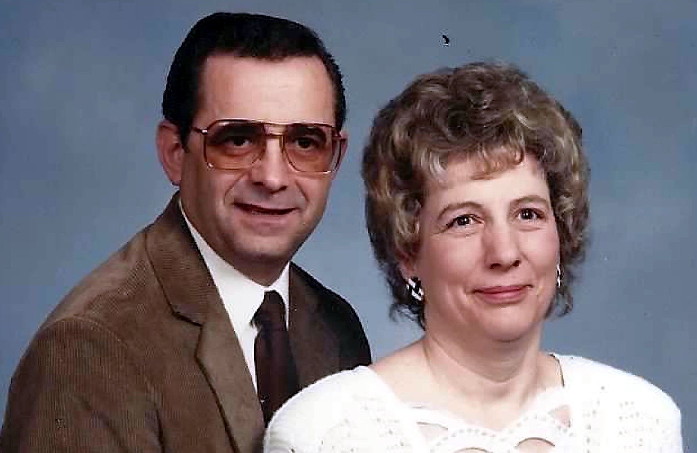Portrait of a middle-aged couple posing together in formal attire, man with brown suit and glasses, woman in a white dress against a blue backdrop.