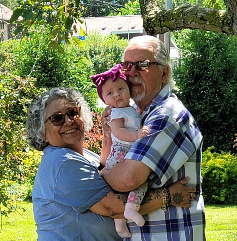 Smiling elderly couple holding a baby girl in a sunlit garden; grandma in blue shirt, grandpa in plaid, baby wears a pink bow headband.