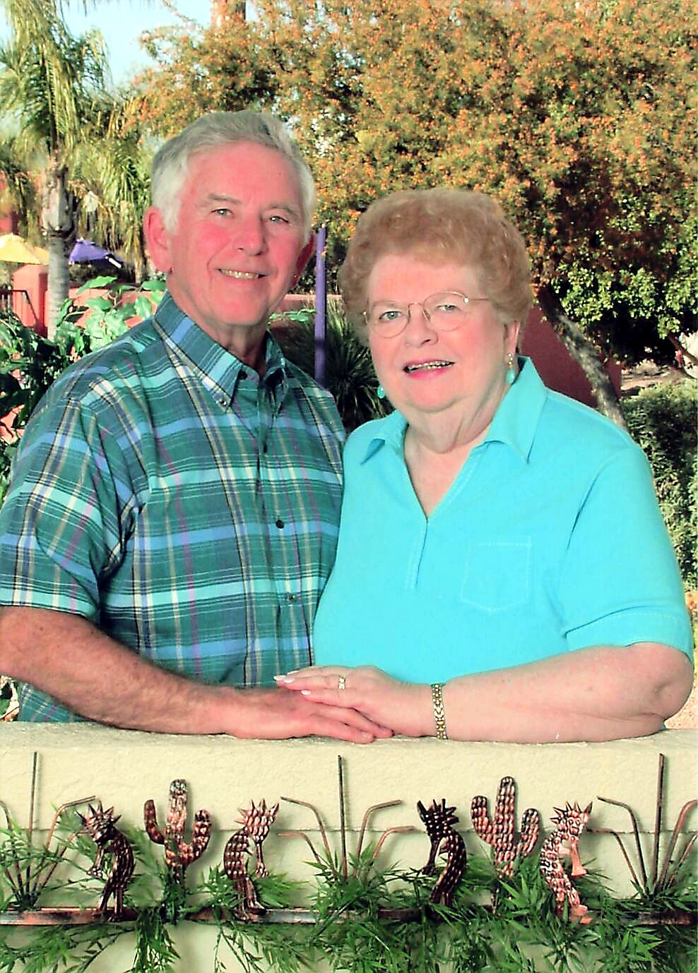 Older couple posing outdoors, hands resting on a low wall with cactus decorations in front of them, smiling at the camera.