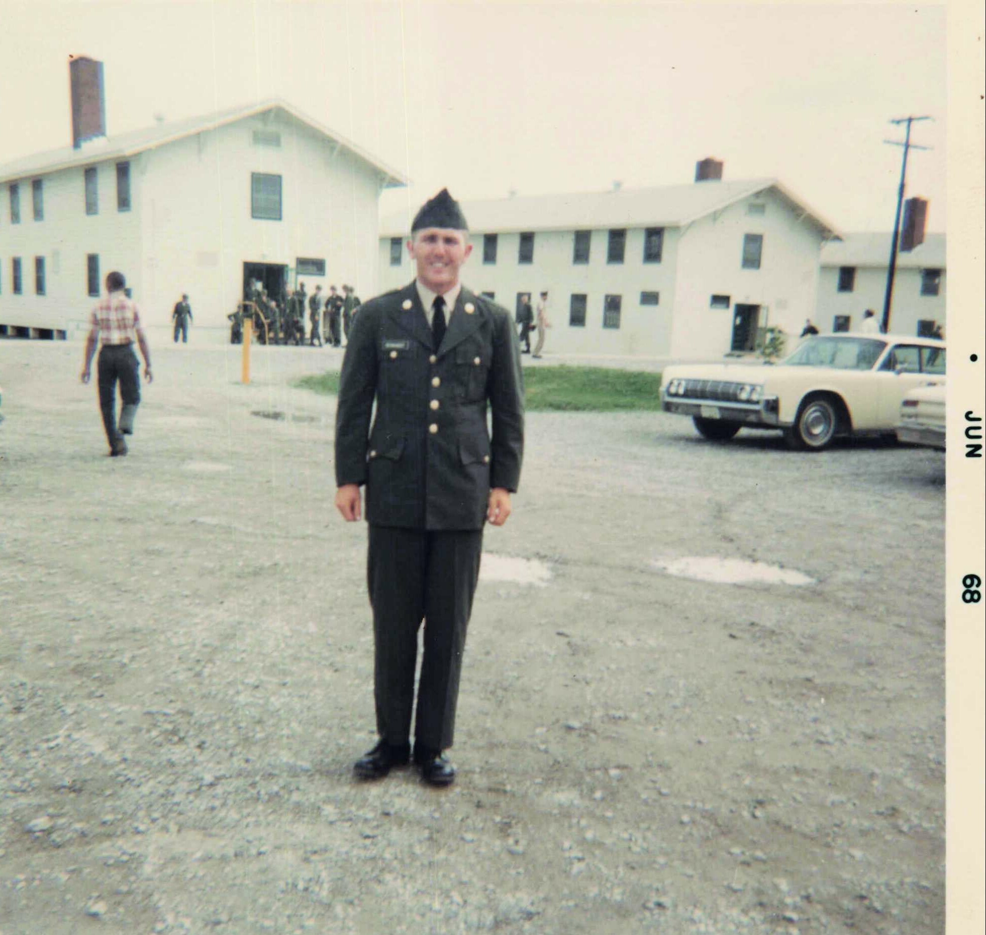 Front-facing man in a formal military uniform standing on a gravel lot with a white barracks-style building behind him.