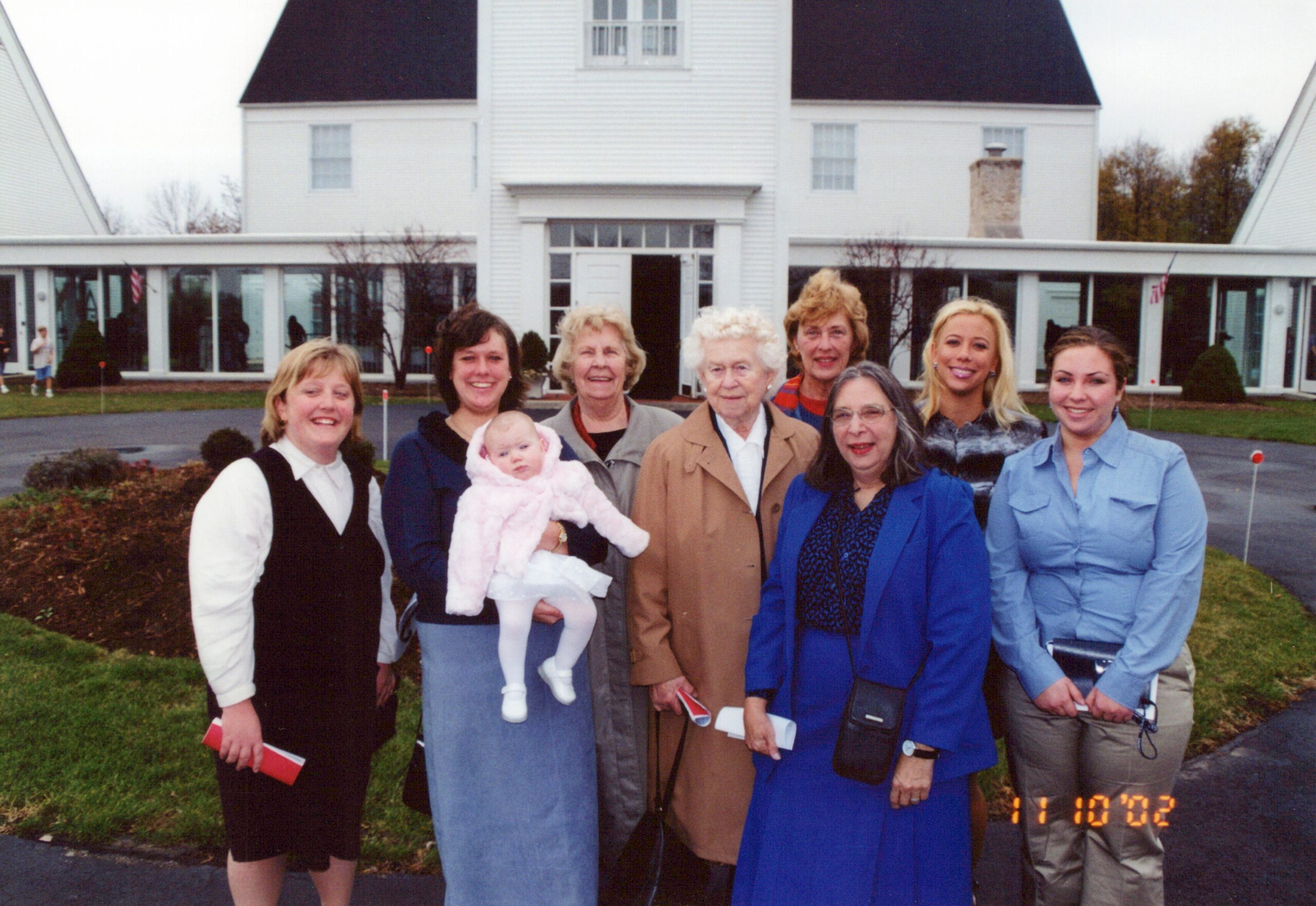 Group of smiling women posing together outside a white building with large windows; one woman holds a baby in a pink outfit.