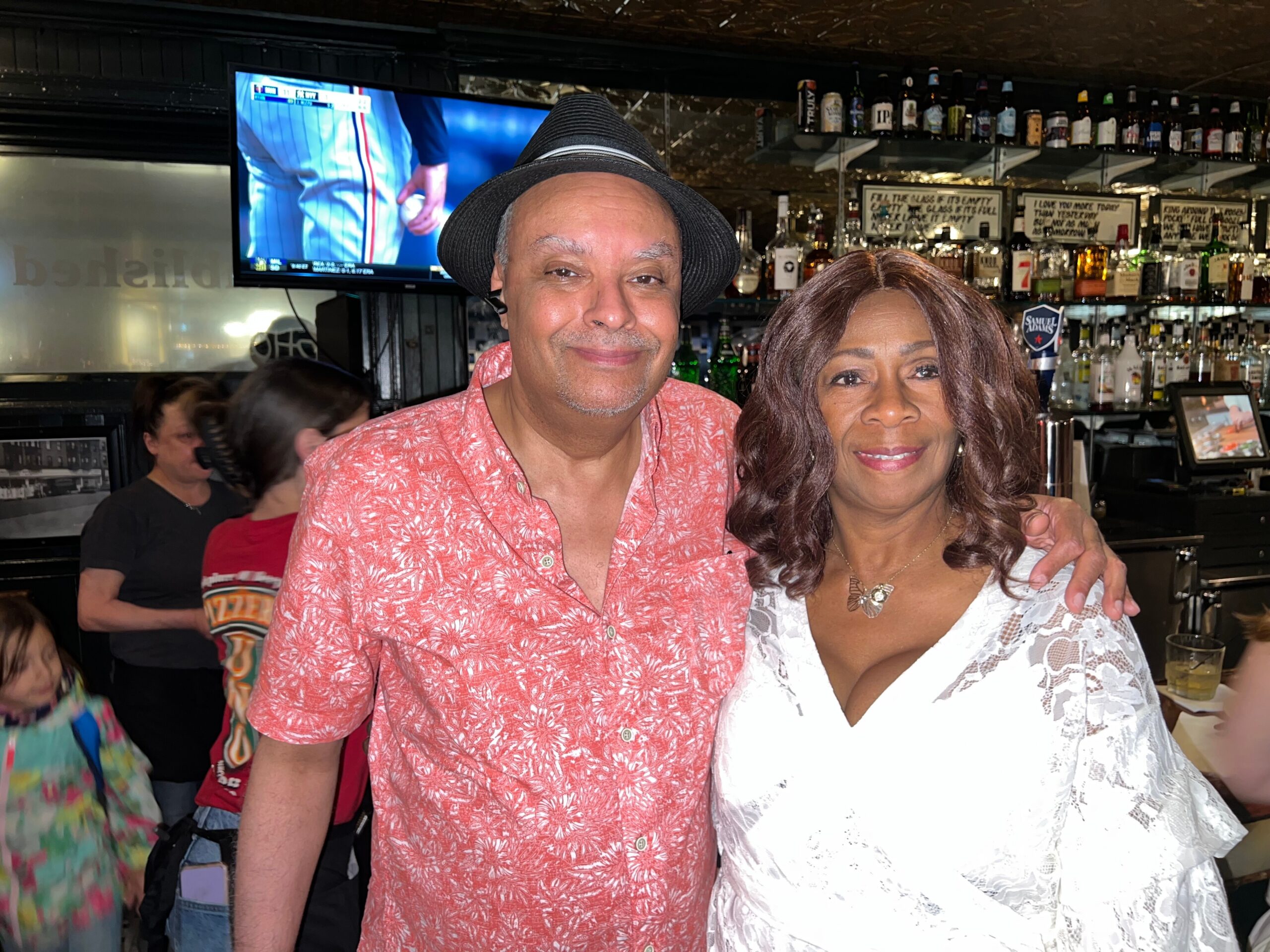 Smiling couple posing at a bar: man in a black hat and pink floral shirt, woman in white lace beside him, liquor shelves in the background.