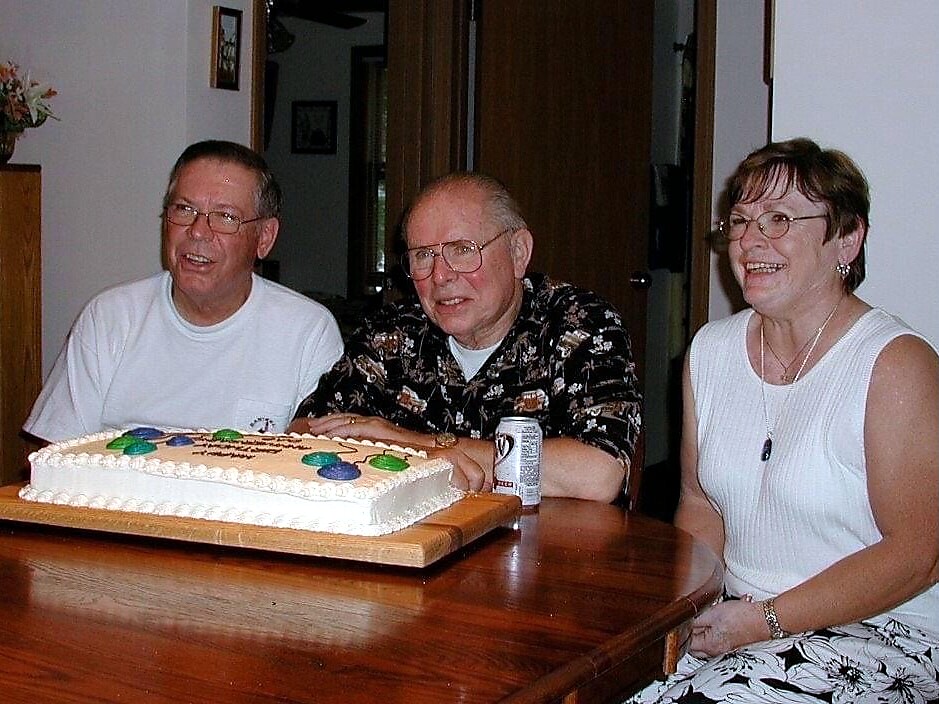 Three adults sit at a kitchen table with a large rectangular cake, smiling for a celebration.