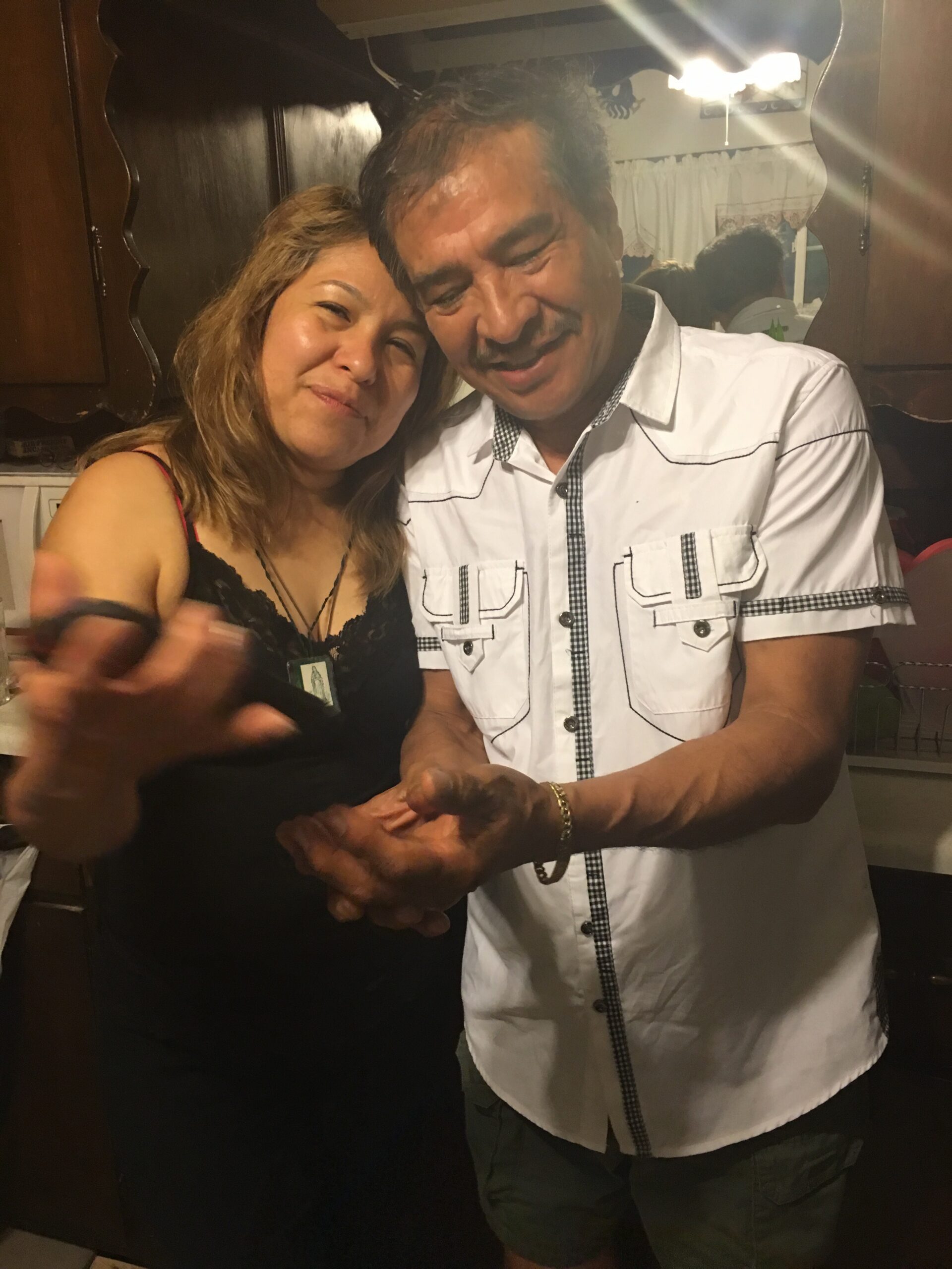 Two adults standing close together, smiling at the camera in a kitchen, with their hands clasped in front.