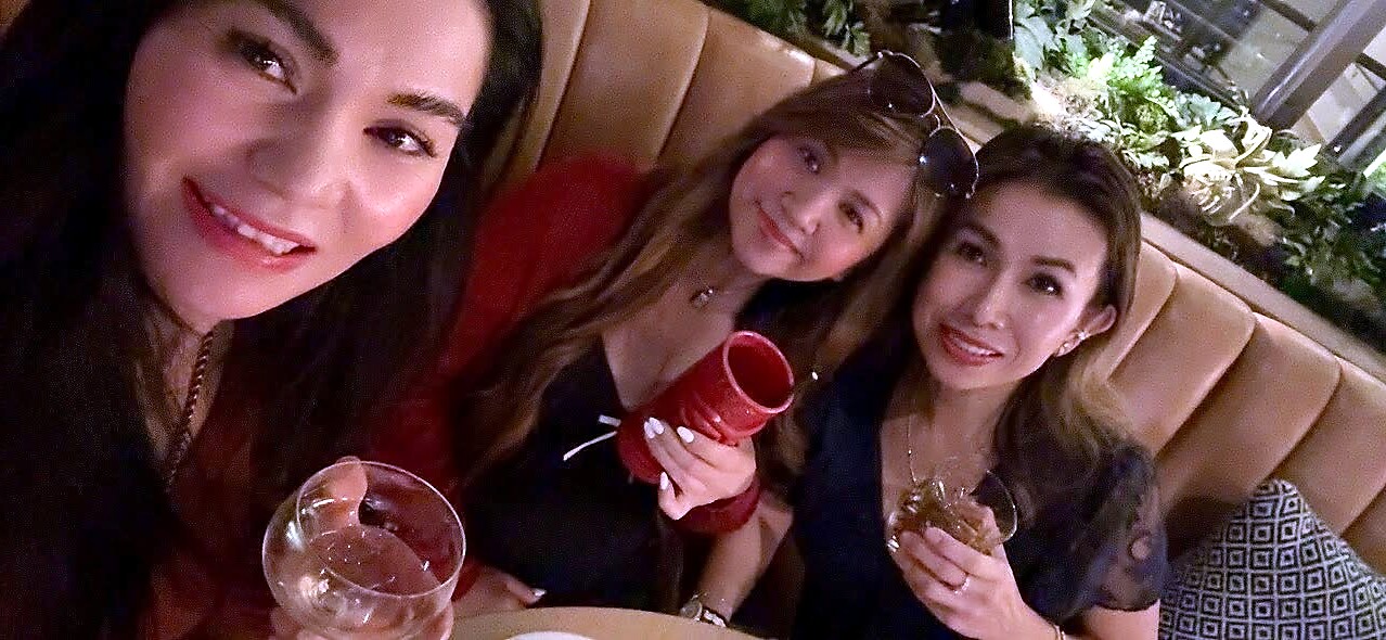 Three women sit close together on a booth, smiling at the camera; one holds a wine glass, one holds a red cup, and the third holds a small plate of food in a restaurant setting with greenery in the background.