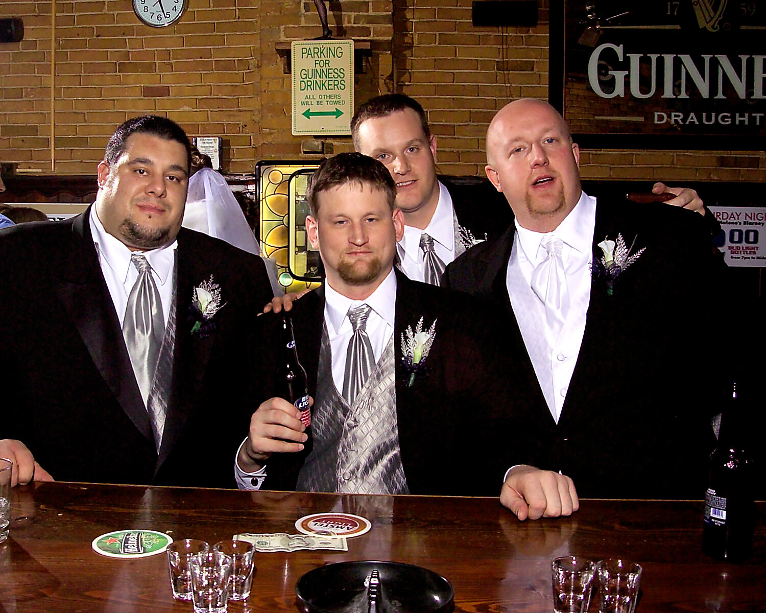 Group of men in suits posing at a bar, one holding a beer bottle, brick wall background.