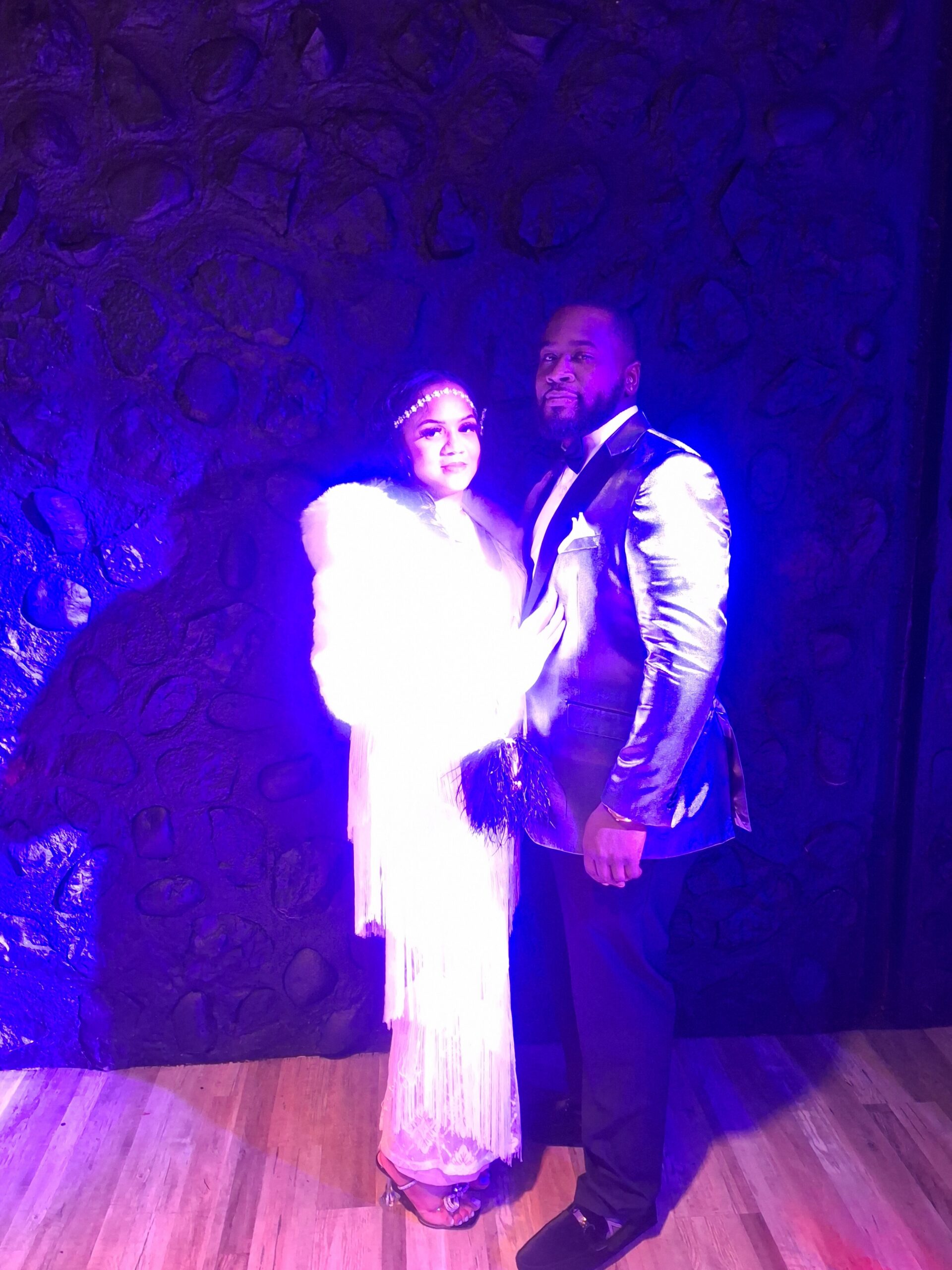 Couple posing in formal attire under blue-purple lighting, standing side by side against a stone wall backdrop.