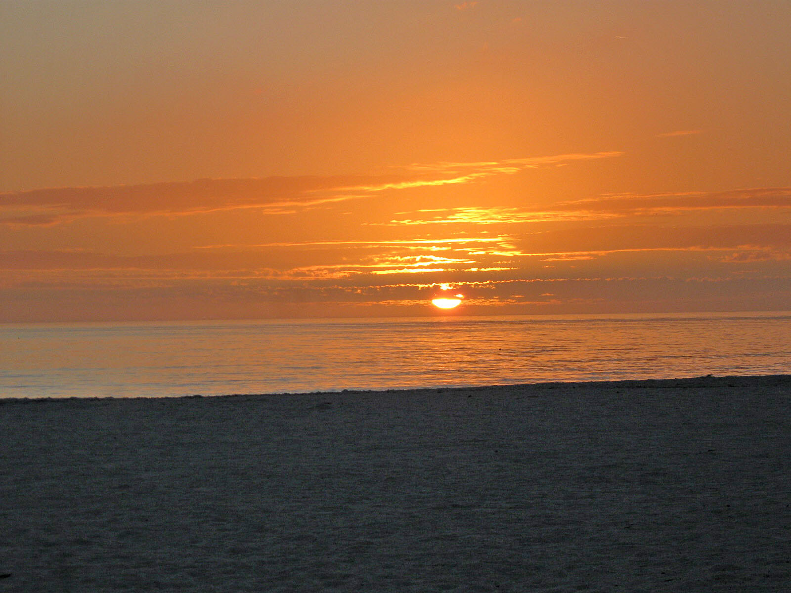 Sunset over the ocean with an orange sky and a darkened sandy beach in the foreground.