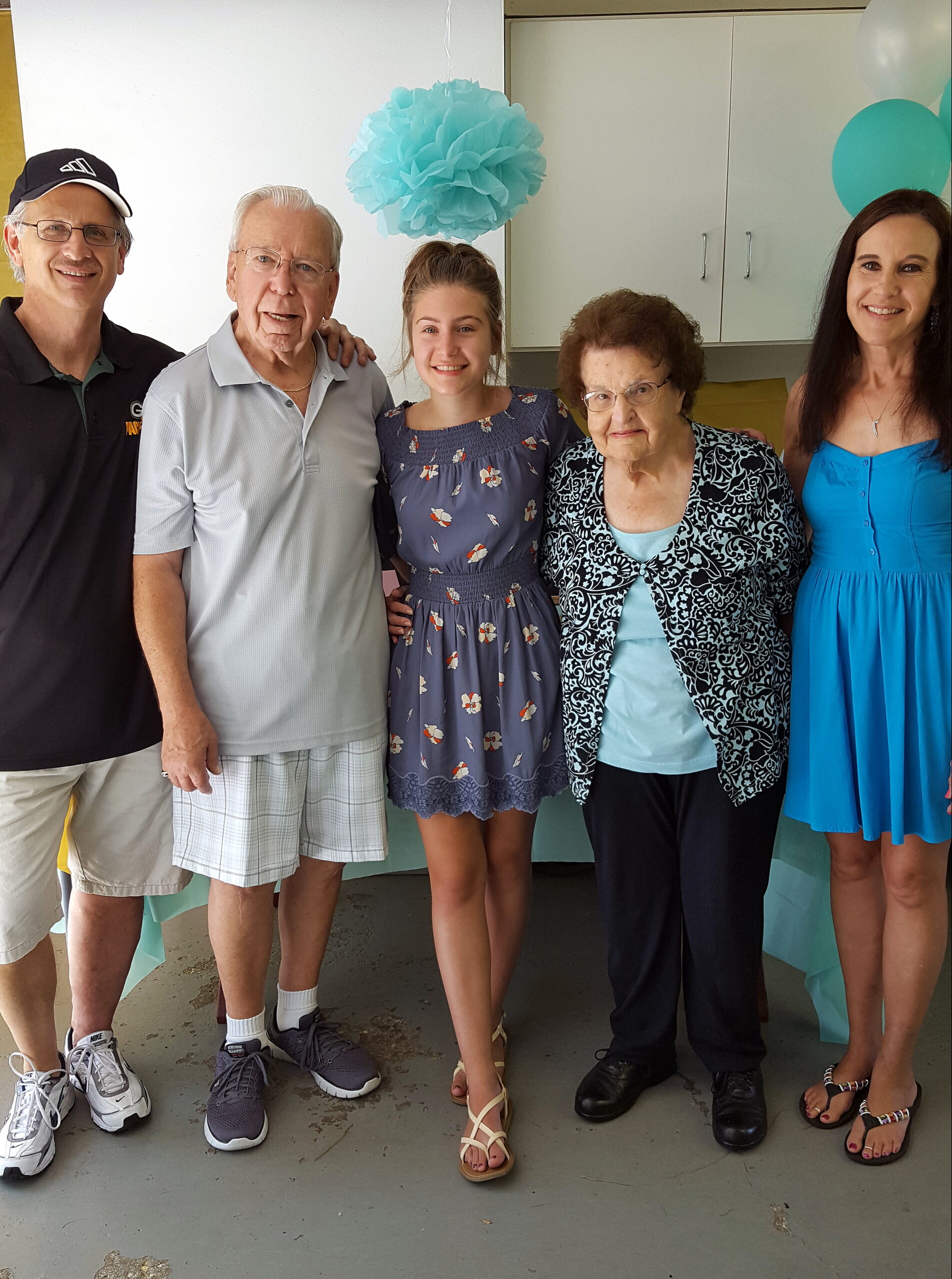 A group of five people posing together indoors, smiling, with blue teal decorations in the background (pom-pom and balloons).