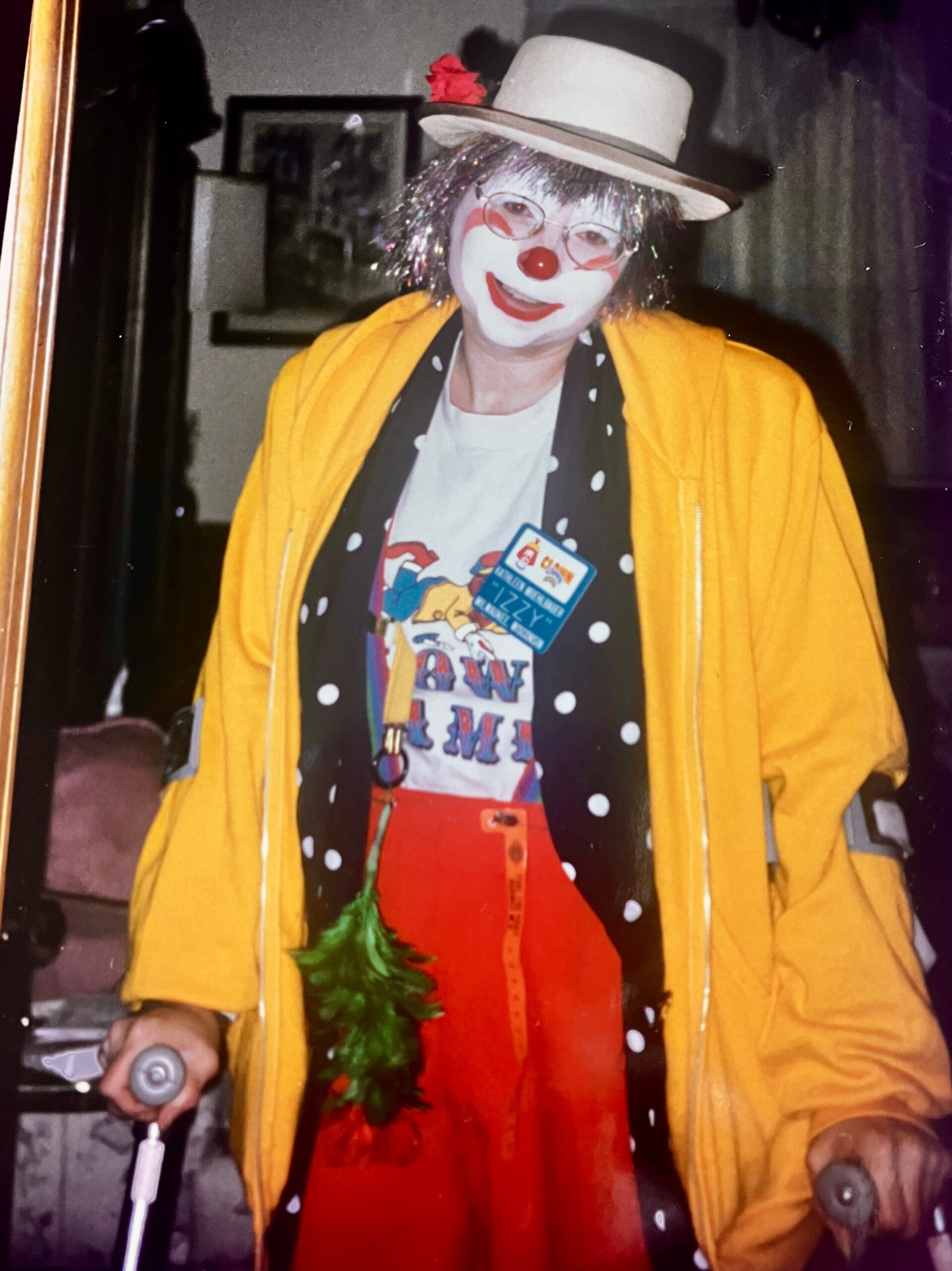 Clown in a yellow coat with white hat, red nose, white makeup, and a polka-dot scarf indoors wearing a name badge.