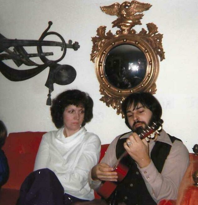 Man with a guitar on a red couch sits beside a woman in a white top; an ornate eagle-motif mirror and a vintage telescope hang on the wall behind them.