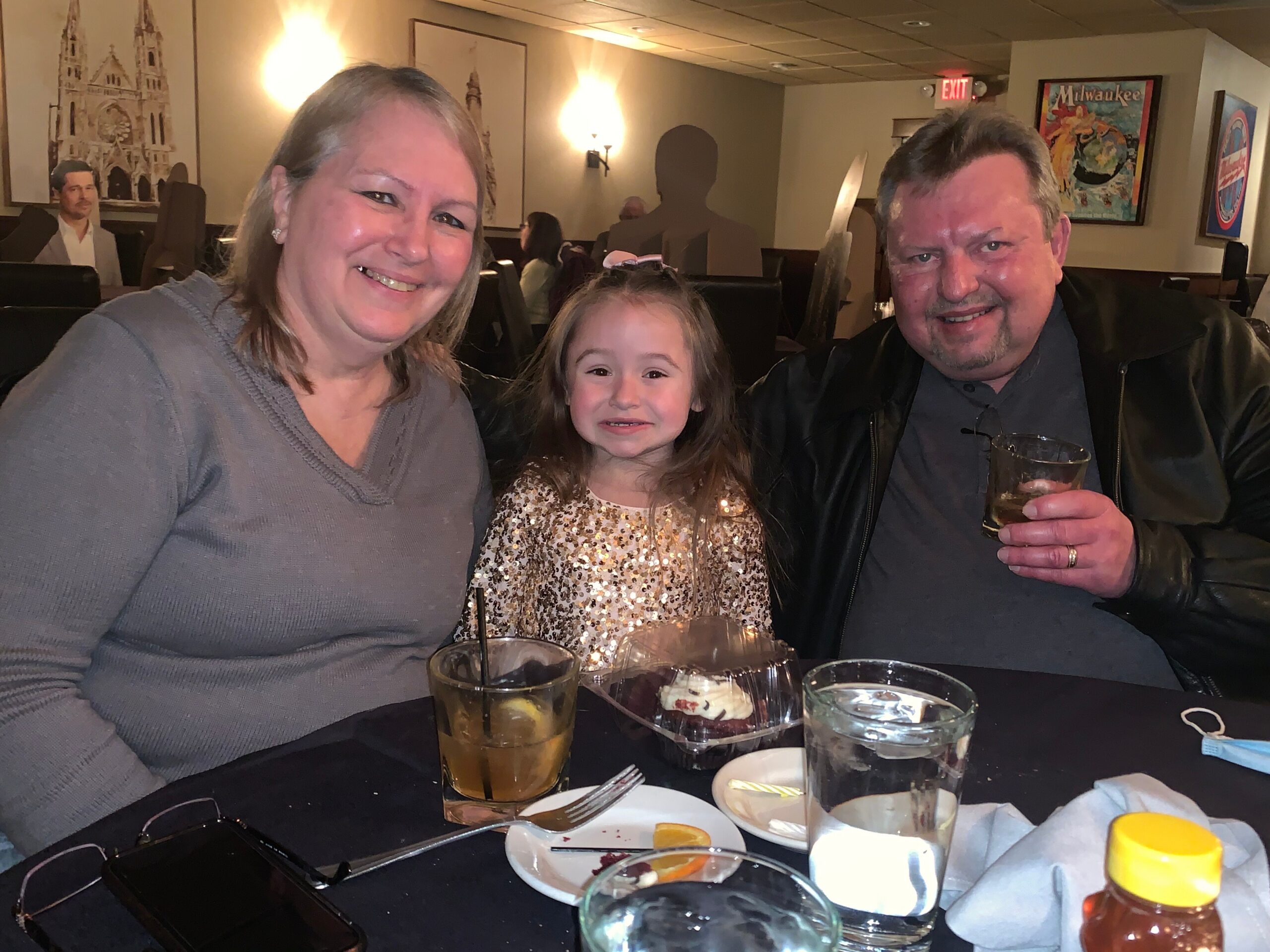 A smiling family of three at a restaurant table: woman on left, young girl center in a sparkly dress, man on right holding a drink.