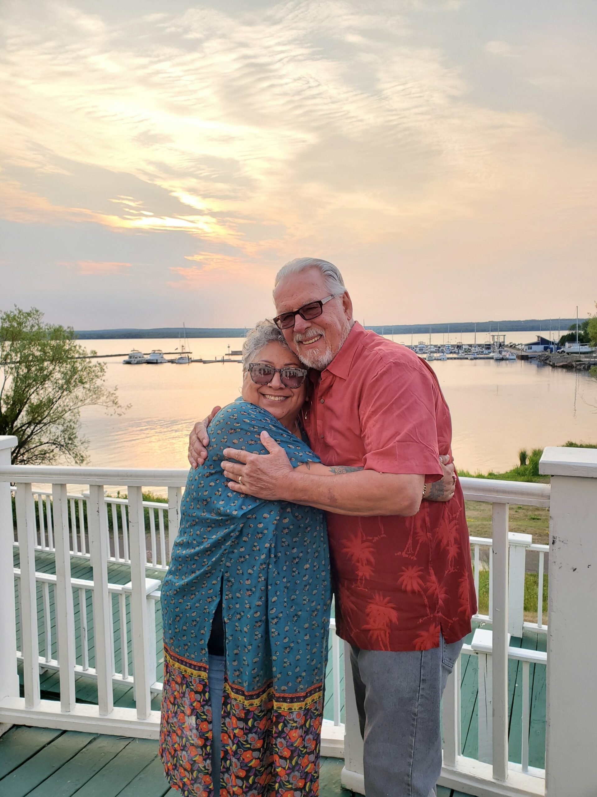 Older couple hugs on a wooden deck by the water at sunset, with boats in a marina in the background, smiling at the camera.