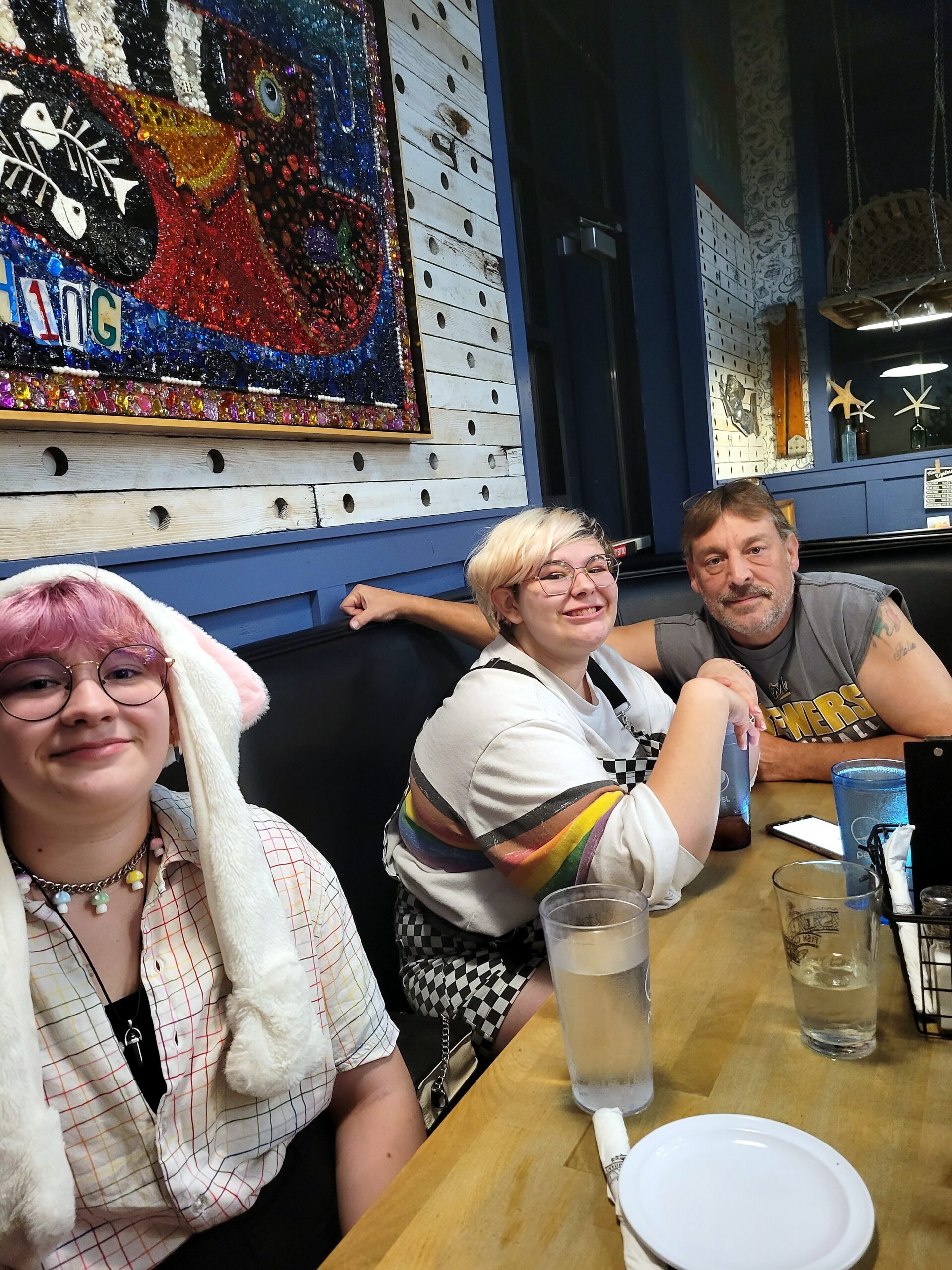 Three friends sit at a wooden table in a restaurant, colorful mural on the wall behind them and drinks in front of them.