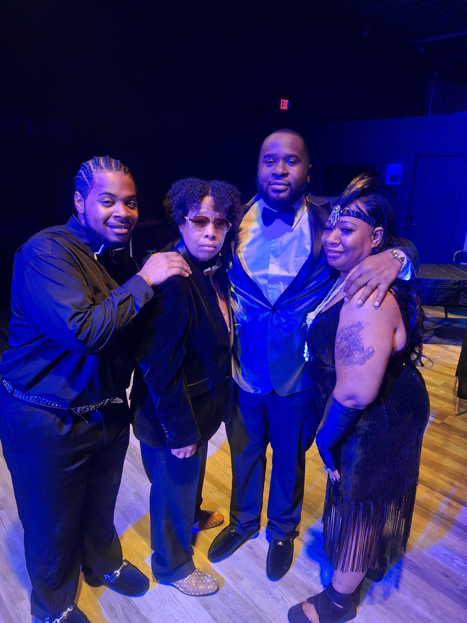 Four friends in formal attire pose together on a stage under blue lighting.