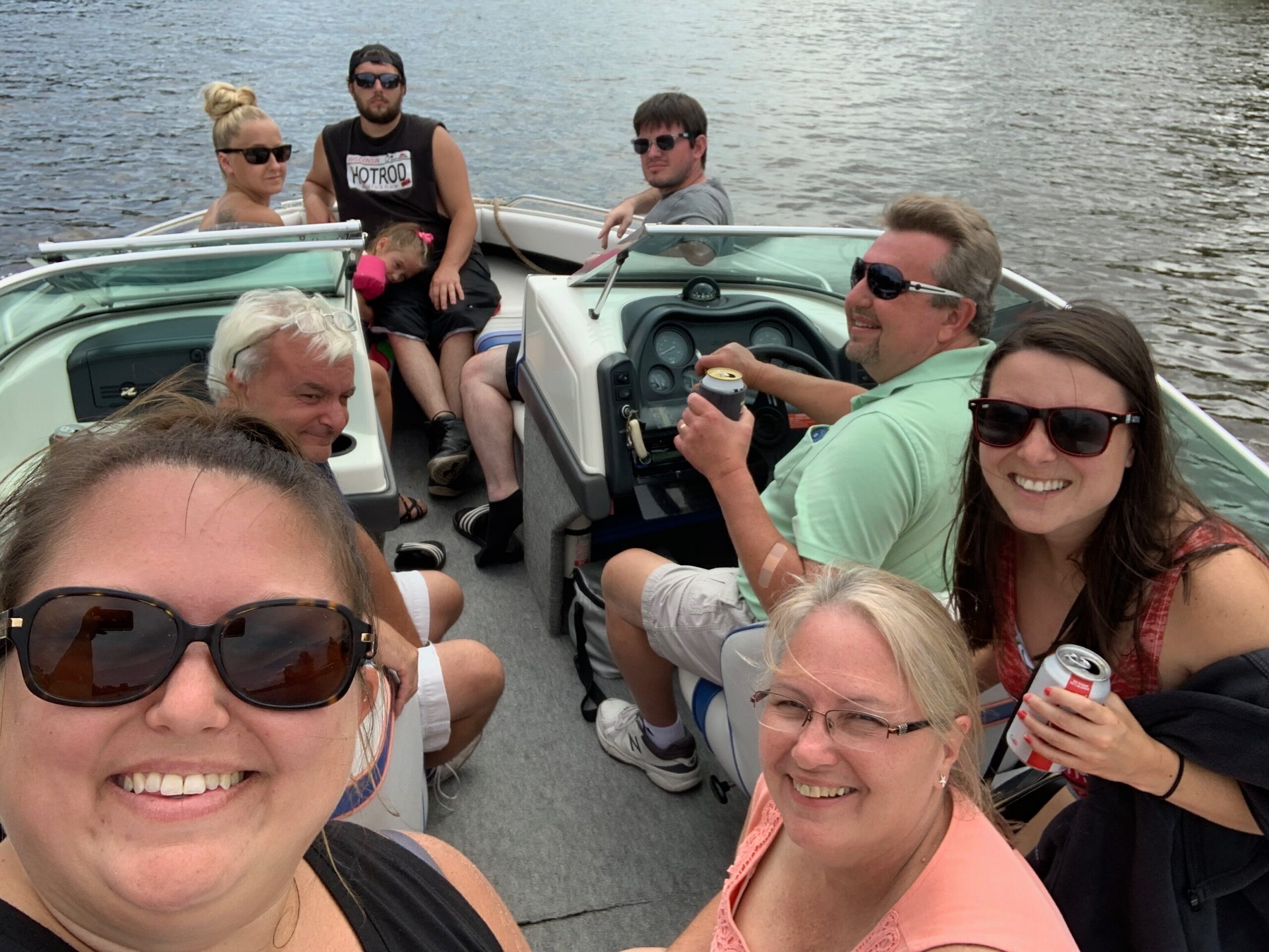 Group of friends on a motorboat taking a sunny selfie, with water in the background and everyone wearing sunglasses.