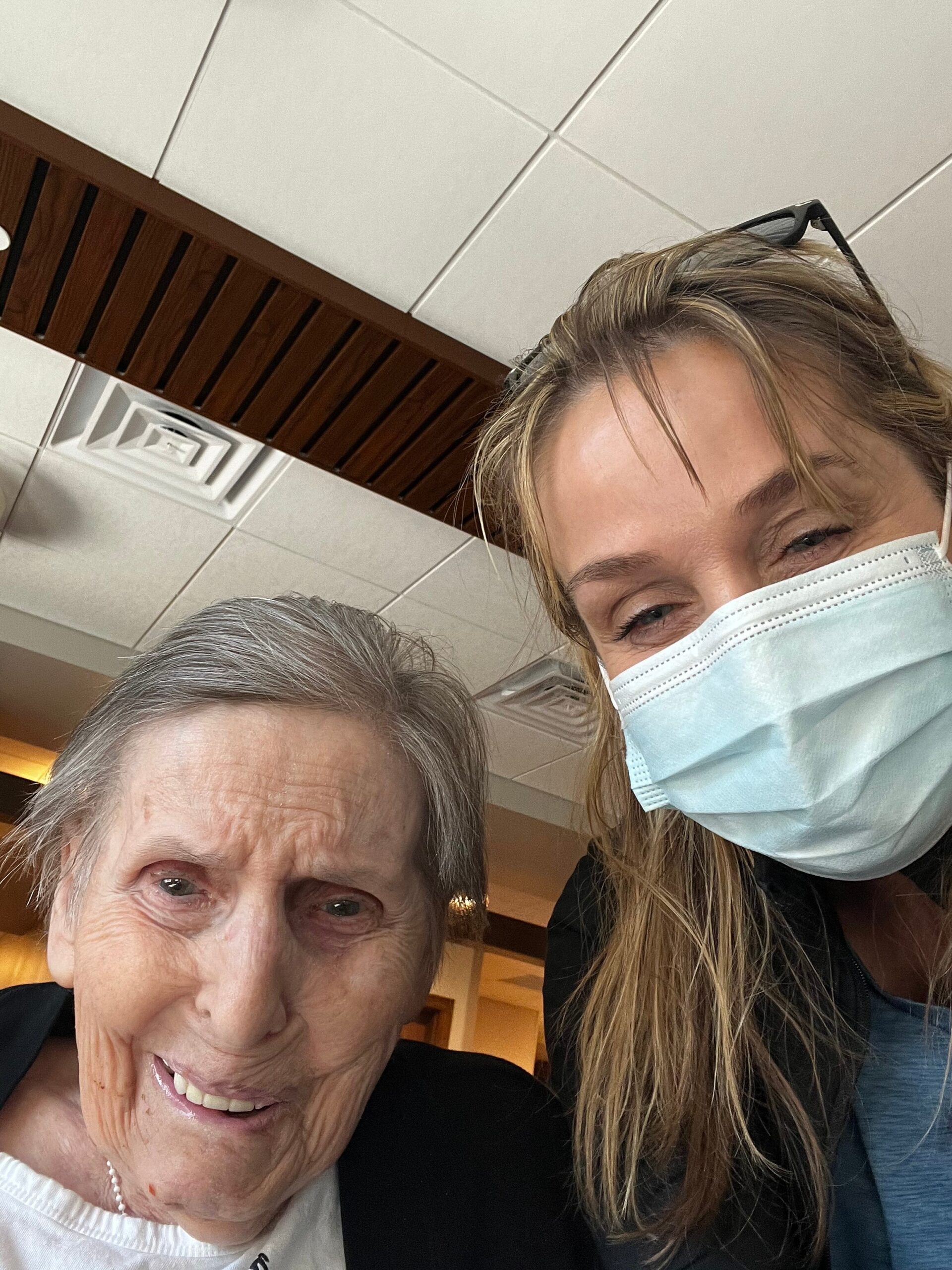 Two women take a close-up selfie indoors; older woman smiling at the camera, younger woman wearing a face mask and sunglasses on her head beside her.