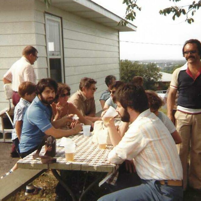 Group of men seated around a checkered picnic table outside a house, chatting and drinking beer.