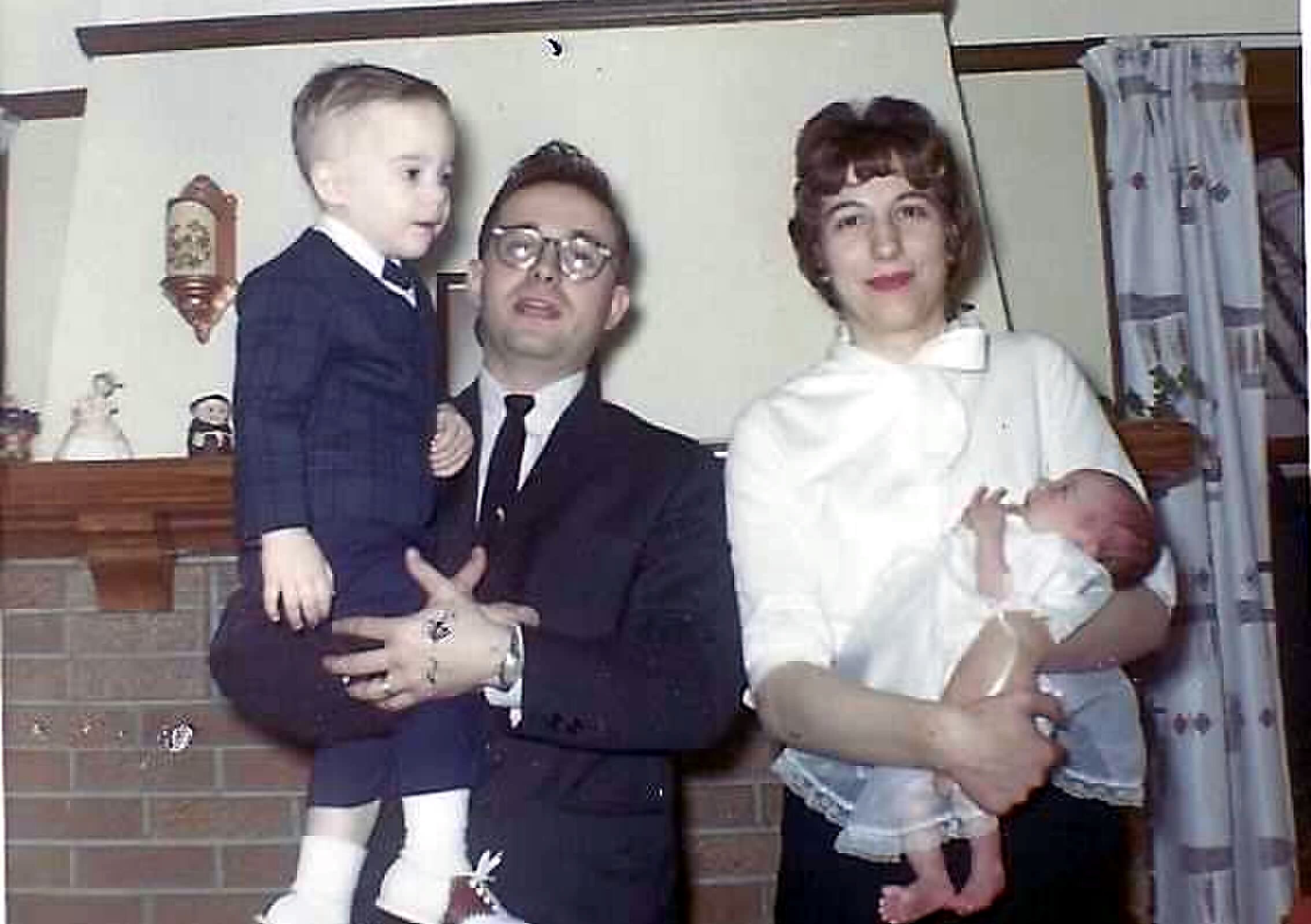 A family portrait indoors: a man in a suit holding a baby, a woman in white holding another baby, with a brick wall and curtains in the background.