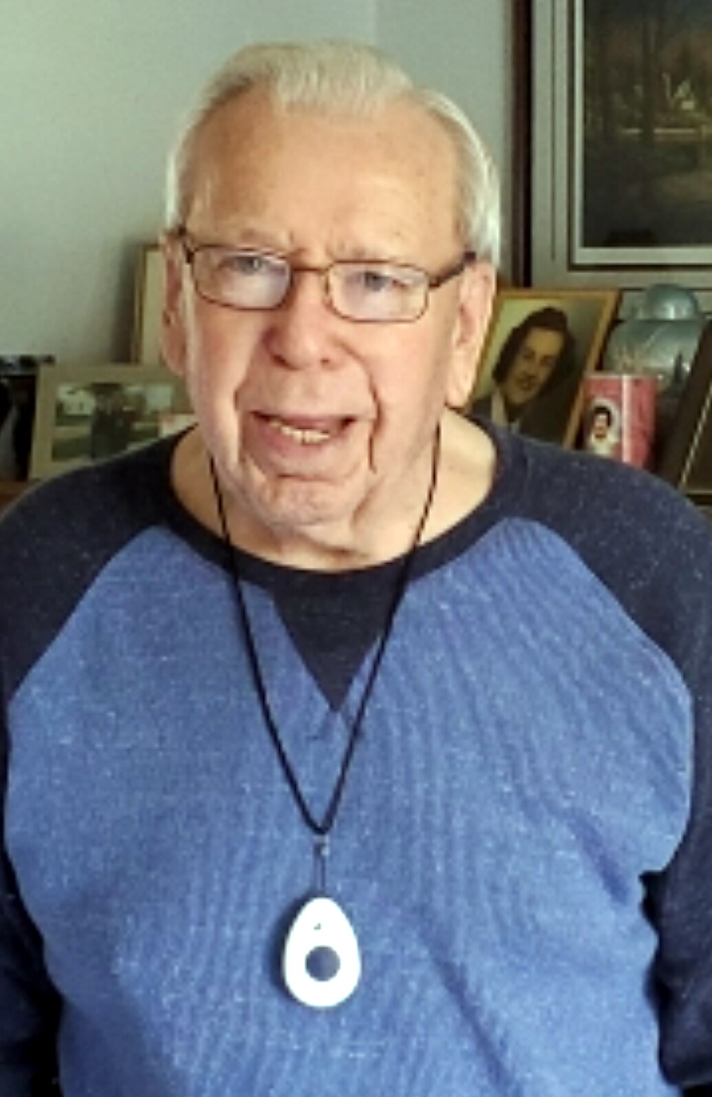 Portrait of an elderly man with white hair and glasses, wearing a blue shirt and a white pendant necklace, smiling at the camera.