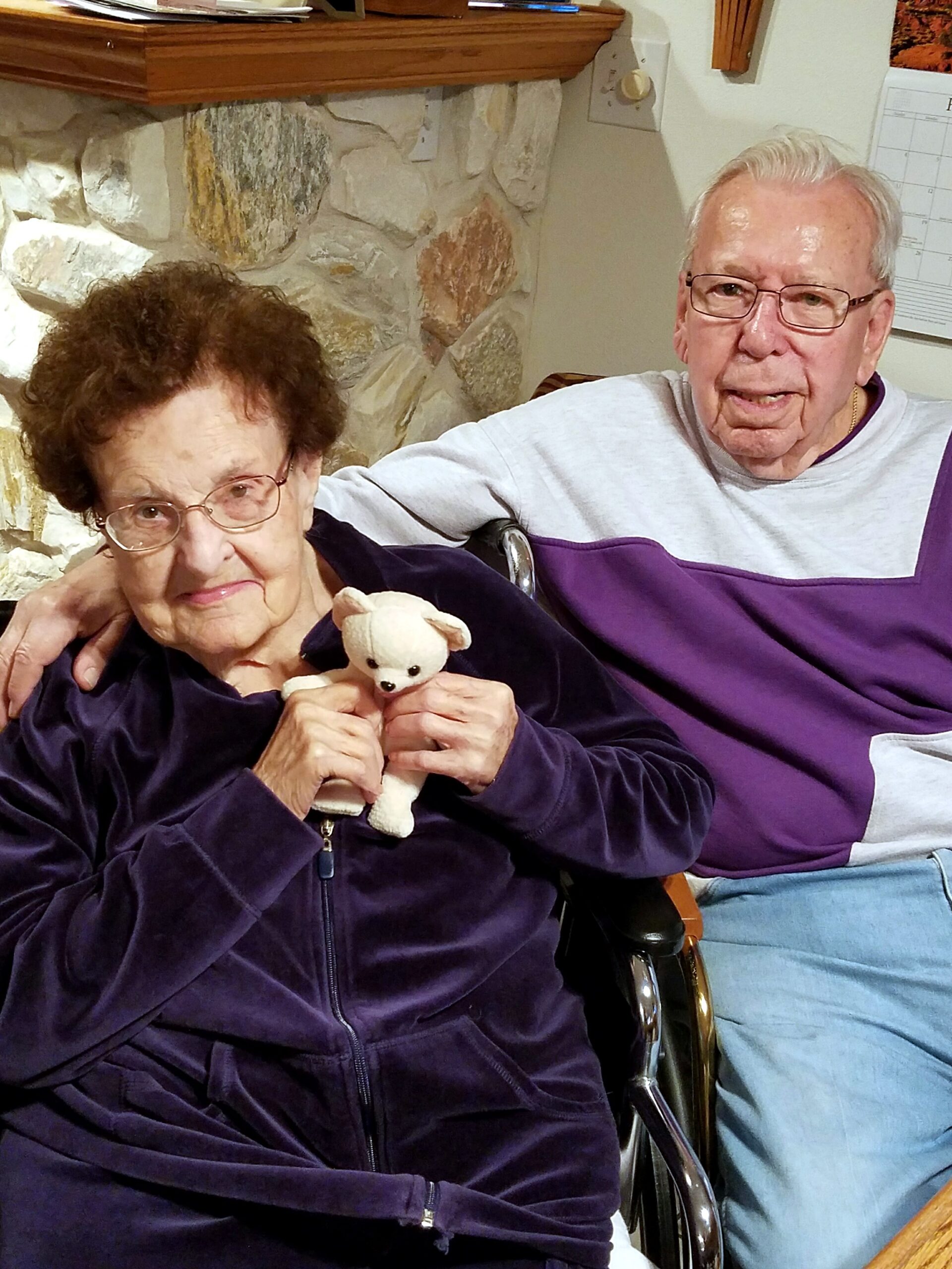Two elderly people sit indoors; woman in a purple velour jacket holds a small white teddy bear, man beside her.