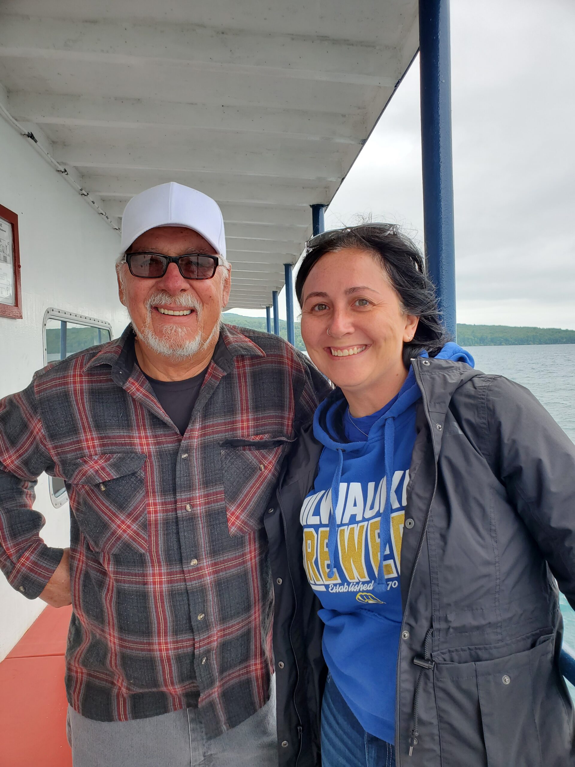 Smiling man in a white cap and plaid shirt and a smiling woman in a blue hoodie stand on a boat deck with water in the background.