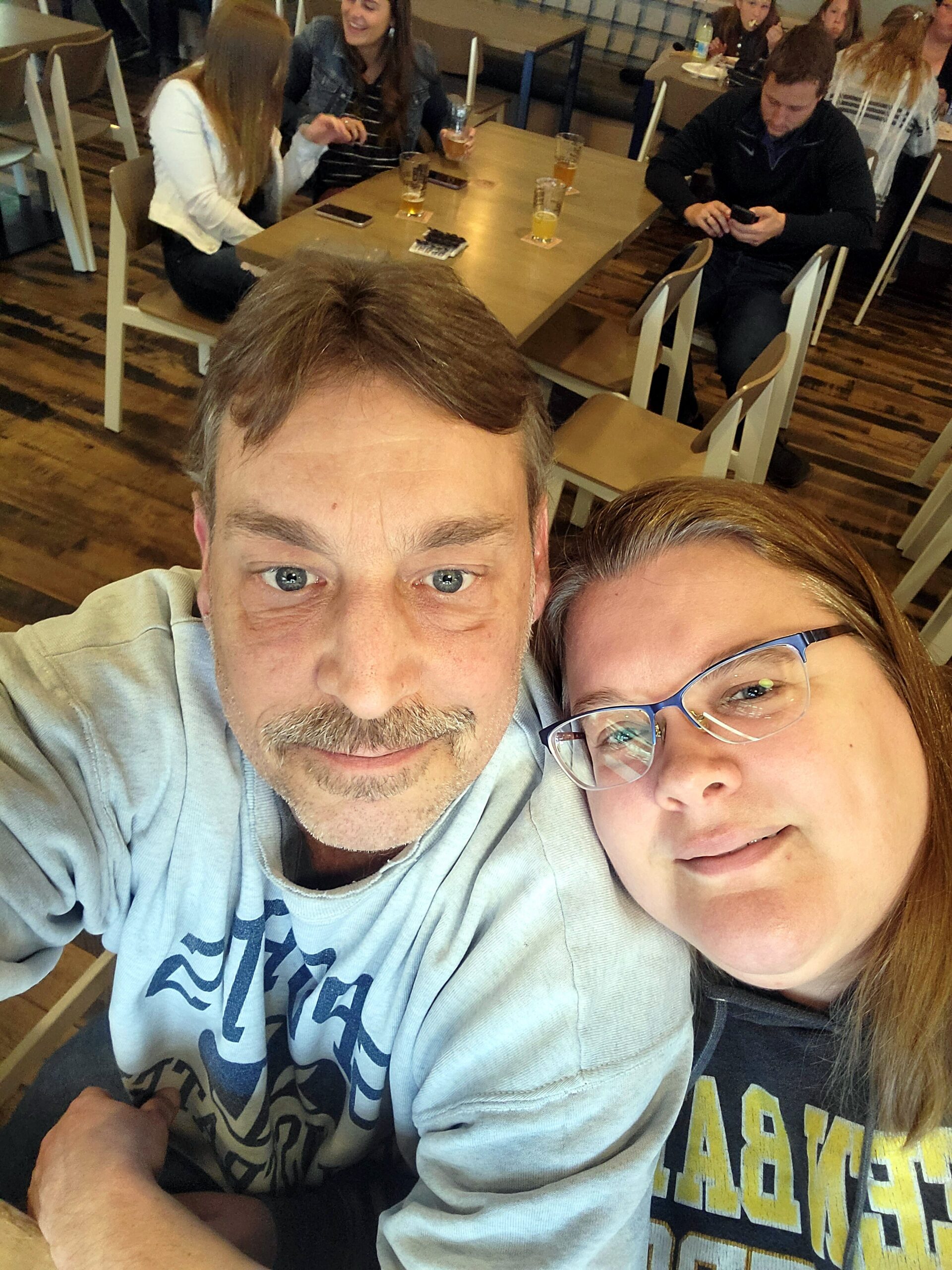 Close-up of a smiling man and woman taking a selfie in a café, with friends and drinks visible at a table in the background.