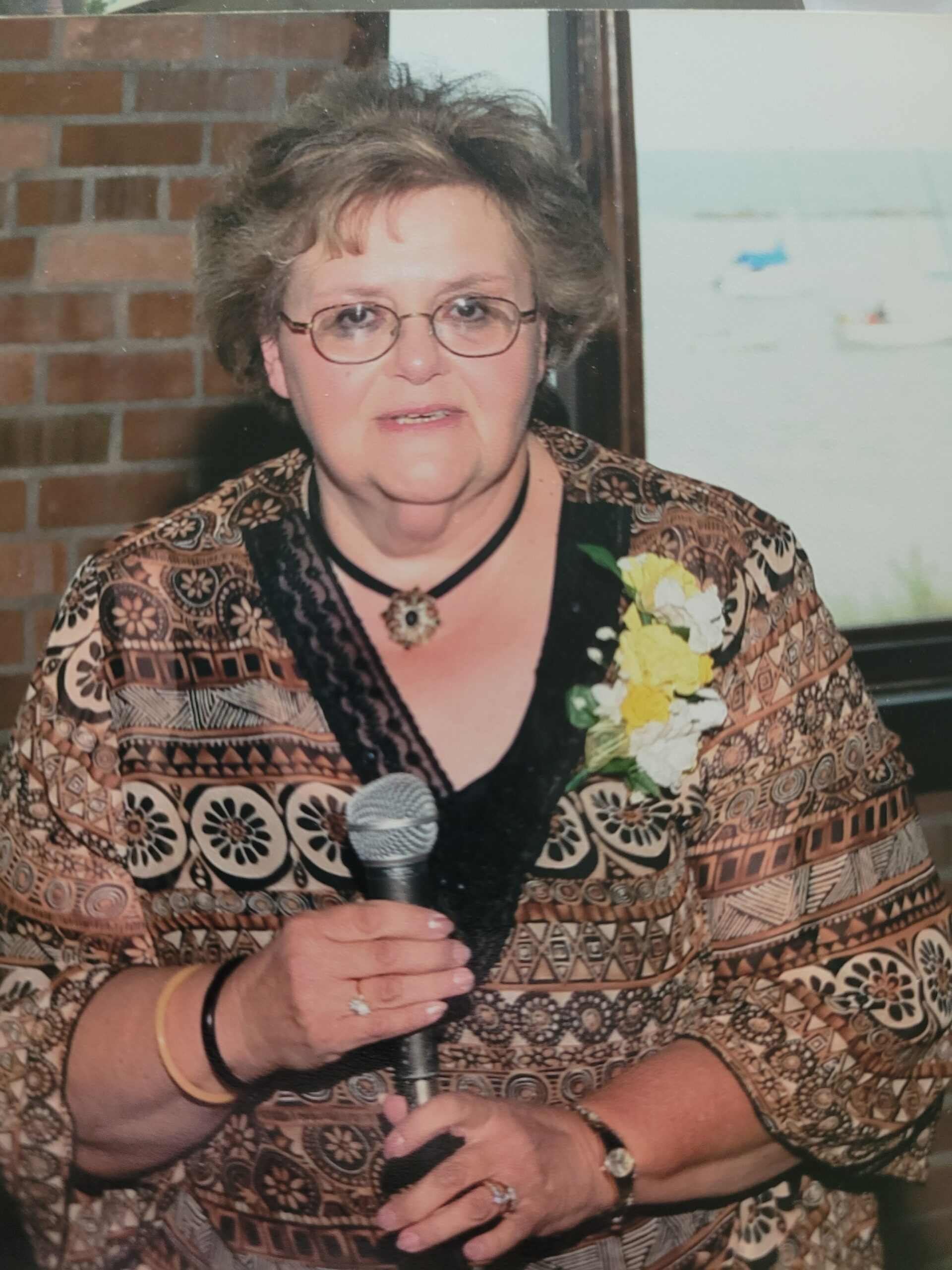 Older woman with glasses wearing a patterned brown jacket, holding a microphone during a speech, with a corsage on her chest.