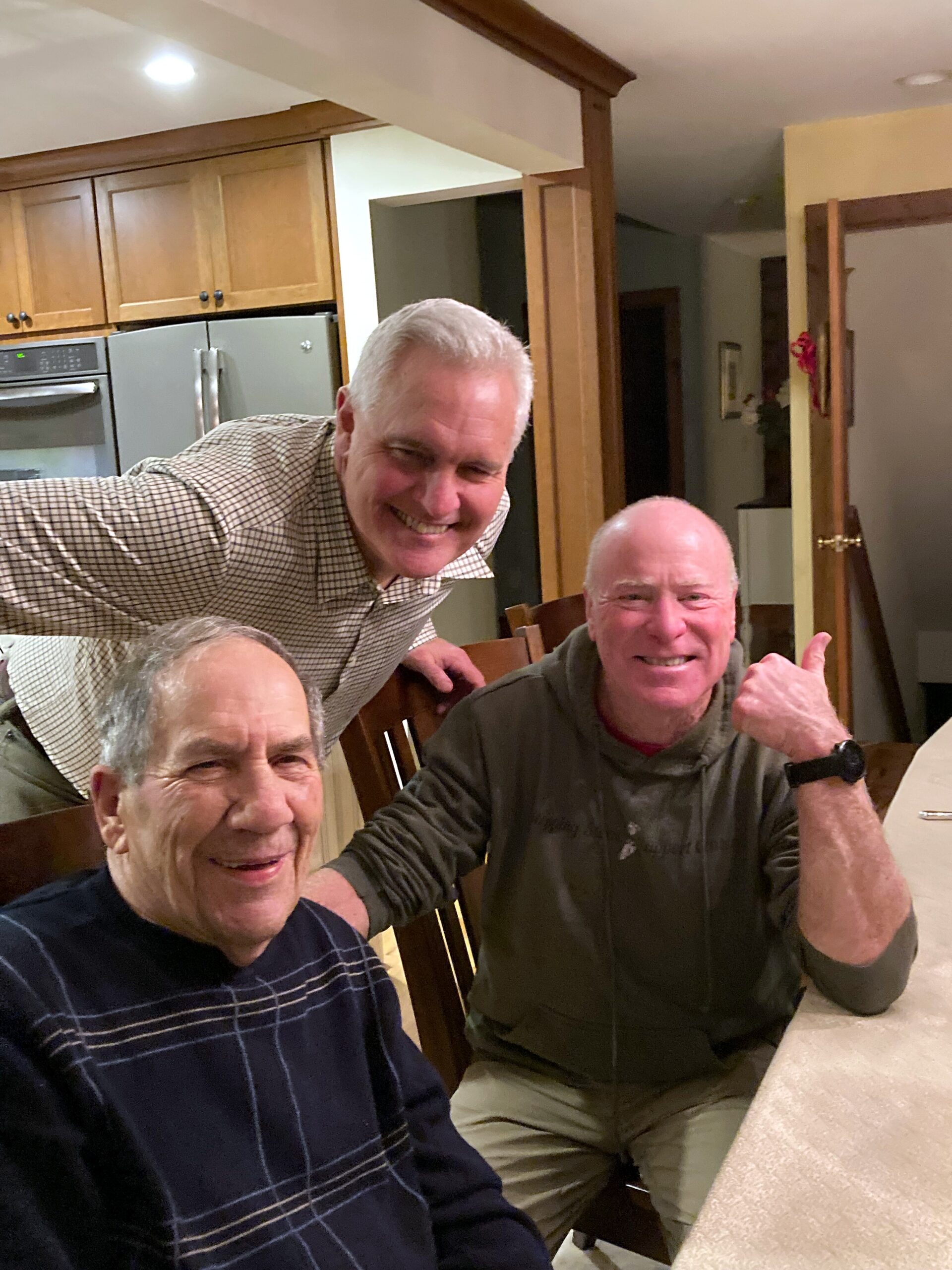 Three smiling older men gathered around a dining table in a home kitchen, posing for a photo.