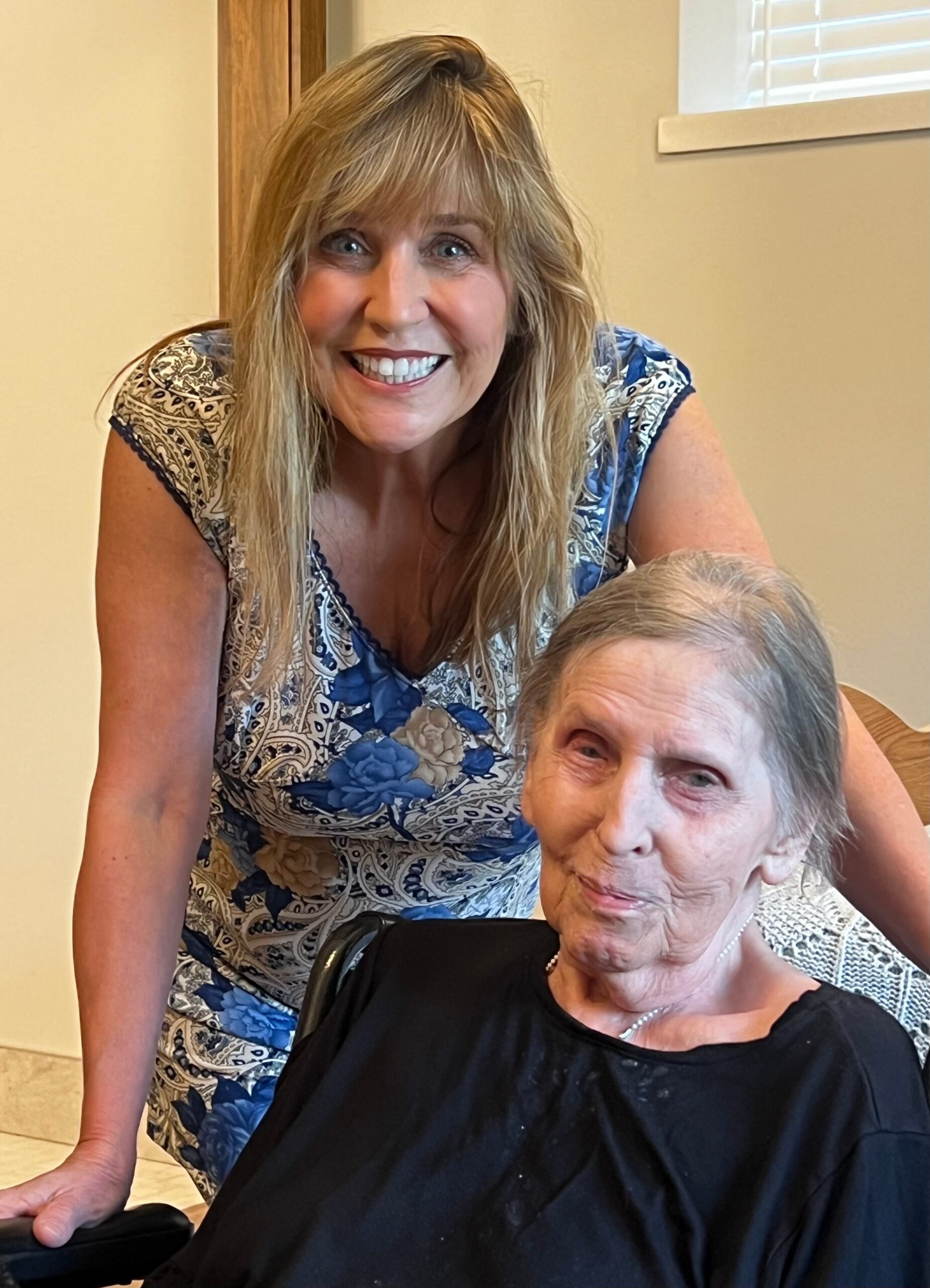 Smiling woman in a blue floral dress leaning over an elderly person seated in a wheelchair indoors.