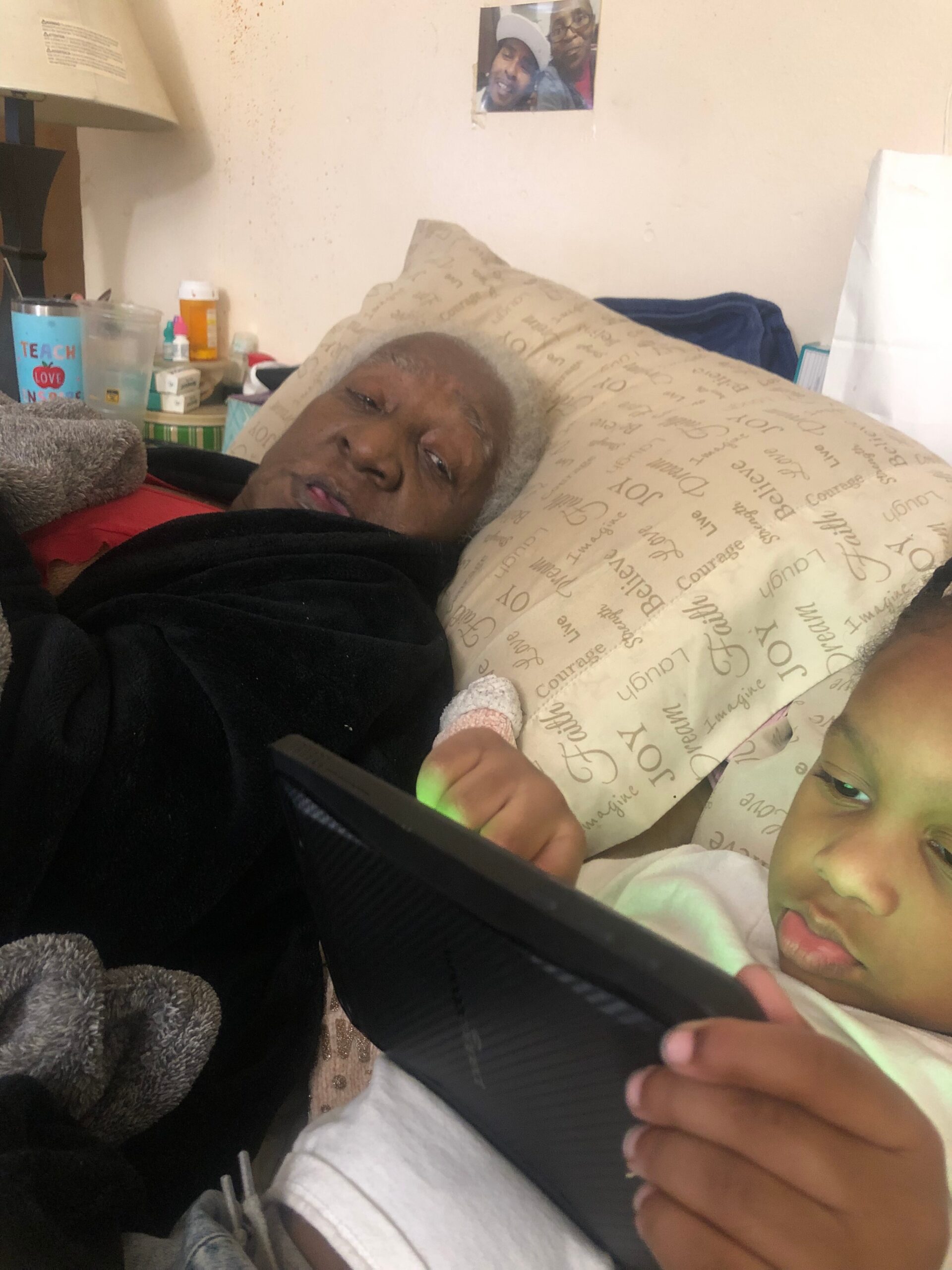 Older woman lying in bed propped by a patterned pillow, with a child beside her touching a black tablet. The room is cluttered with meds and cups in the background.