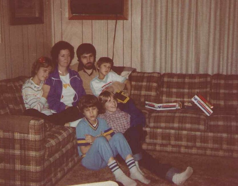 Family of six sitting on a brown plaid sofa in a retro living room, posing for a photo with books on the armrest