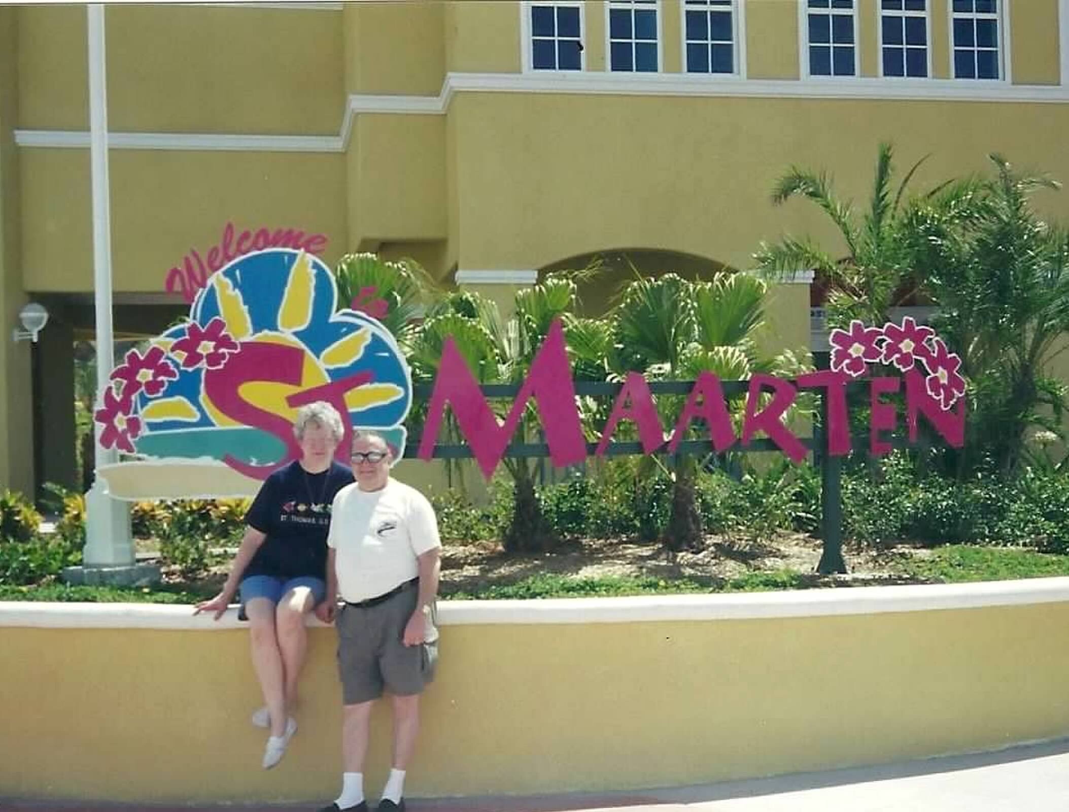 Two seniors sitting on a yellow wall in front of a building with tropical, colorful decorations and plants behind them.