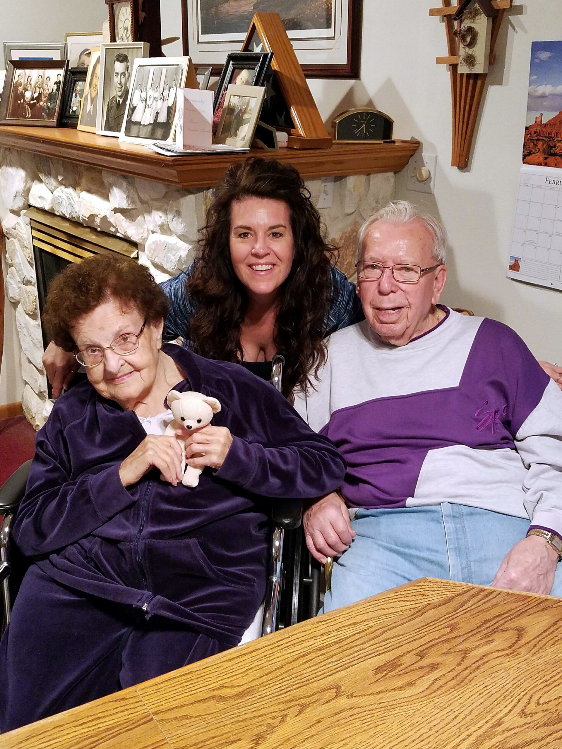 Smiling trio sits on a living room couch, younger woman between an elderly couple; elderly woman holds a teddy bear, stone fireplace in background.