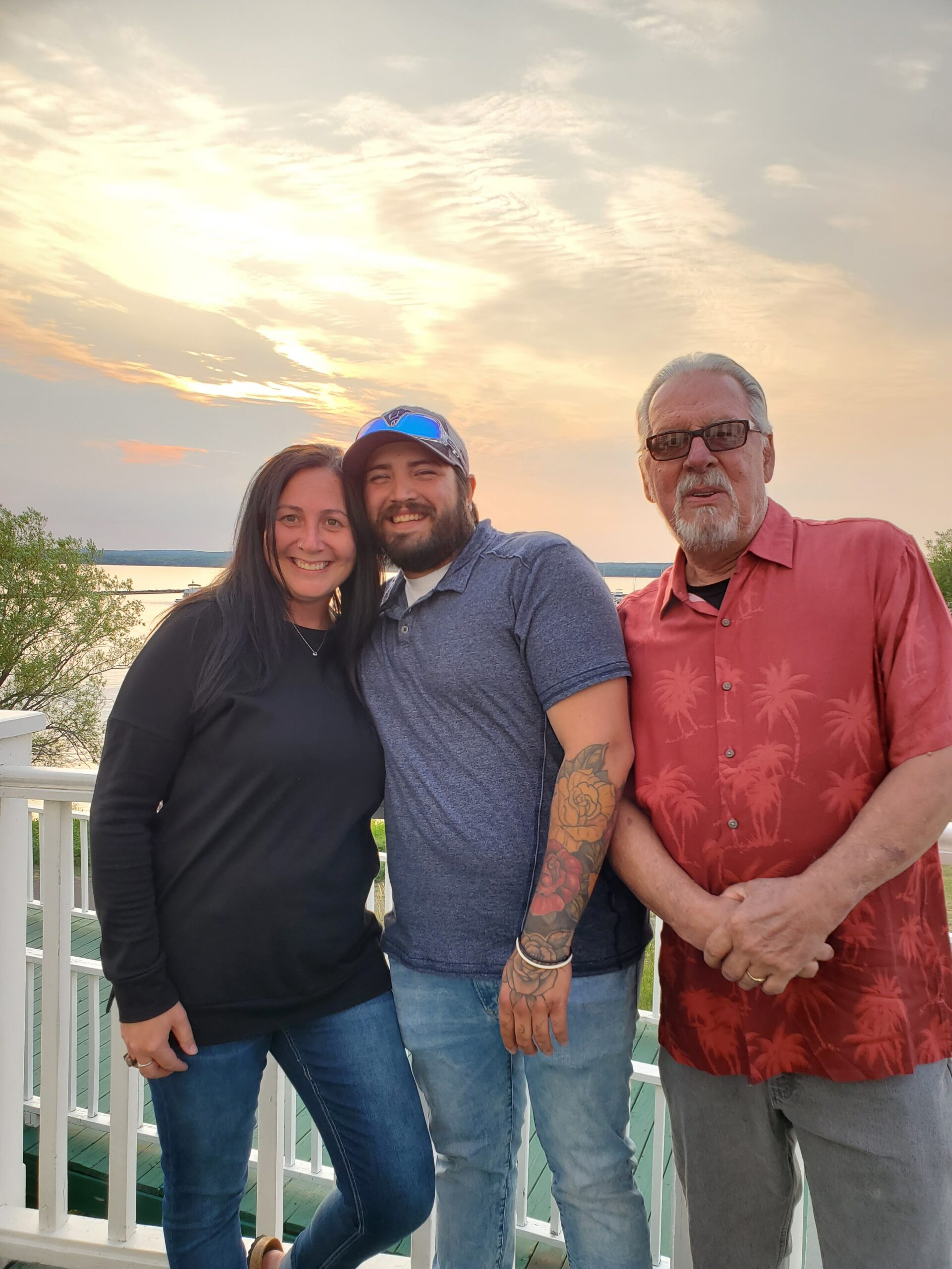 Three adults posing together on a railing at sunset by a lake, smiling at the camera.