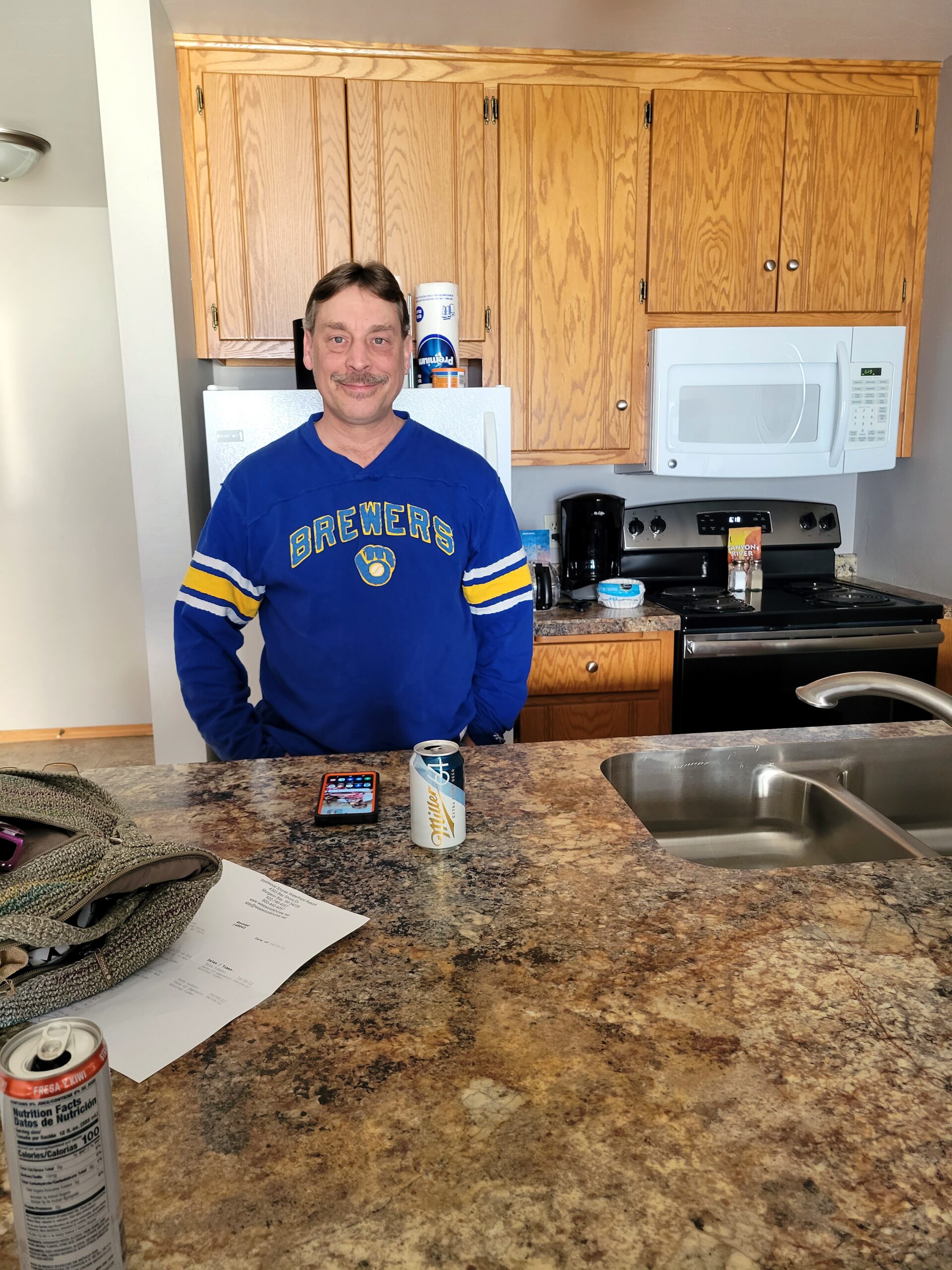 Man in a blue Brewers shirt smiles at the camera in a kitchen with oak cabinets, a white microwave, and a granite countertop.
