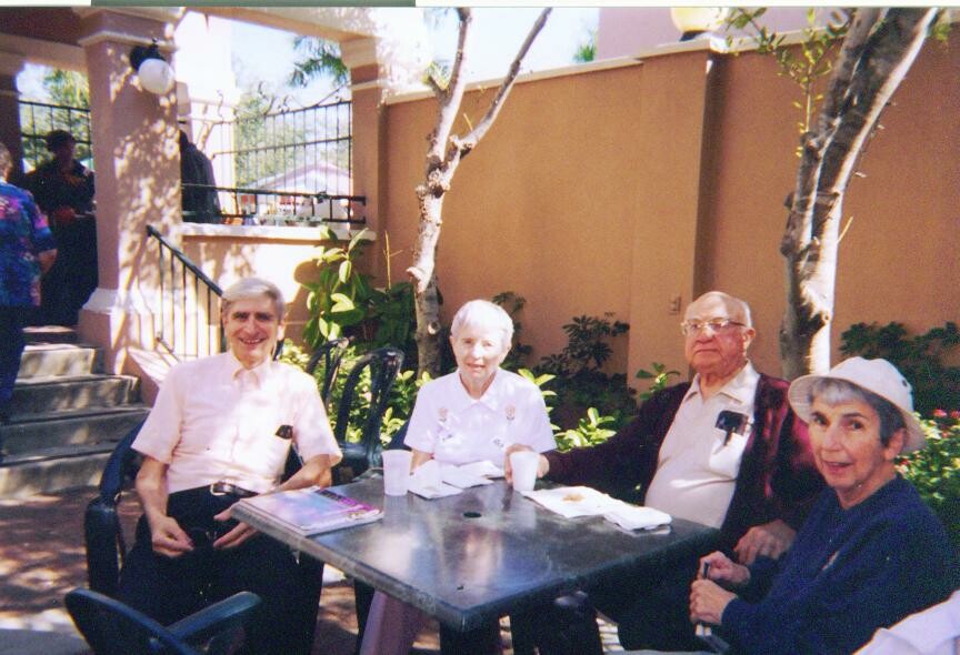 Four older adults sit at a metal table in a sunny courtyard, with drinks and snacks on the table, beneath trees and a beige wall.