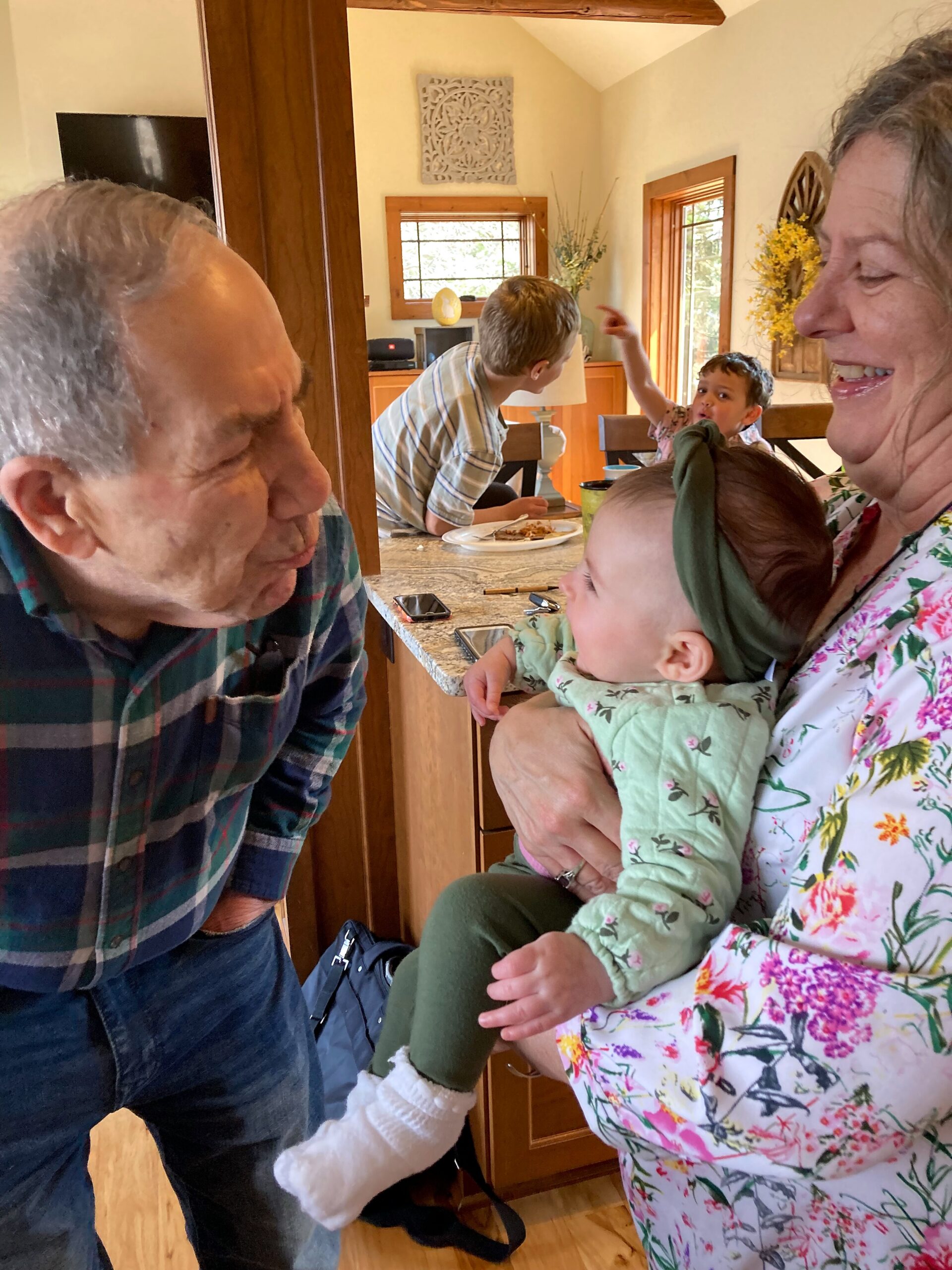 Grandfather leans in to kiss a baby held by a smiling grandmother inside a bright kitchen/dining area.